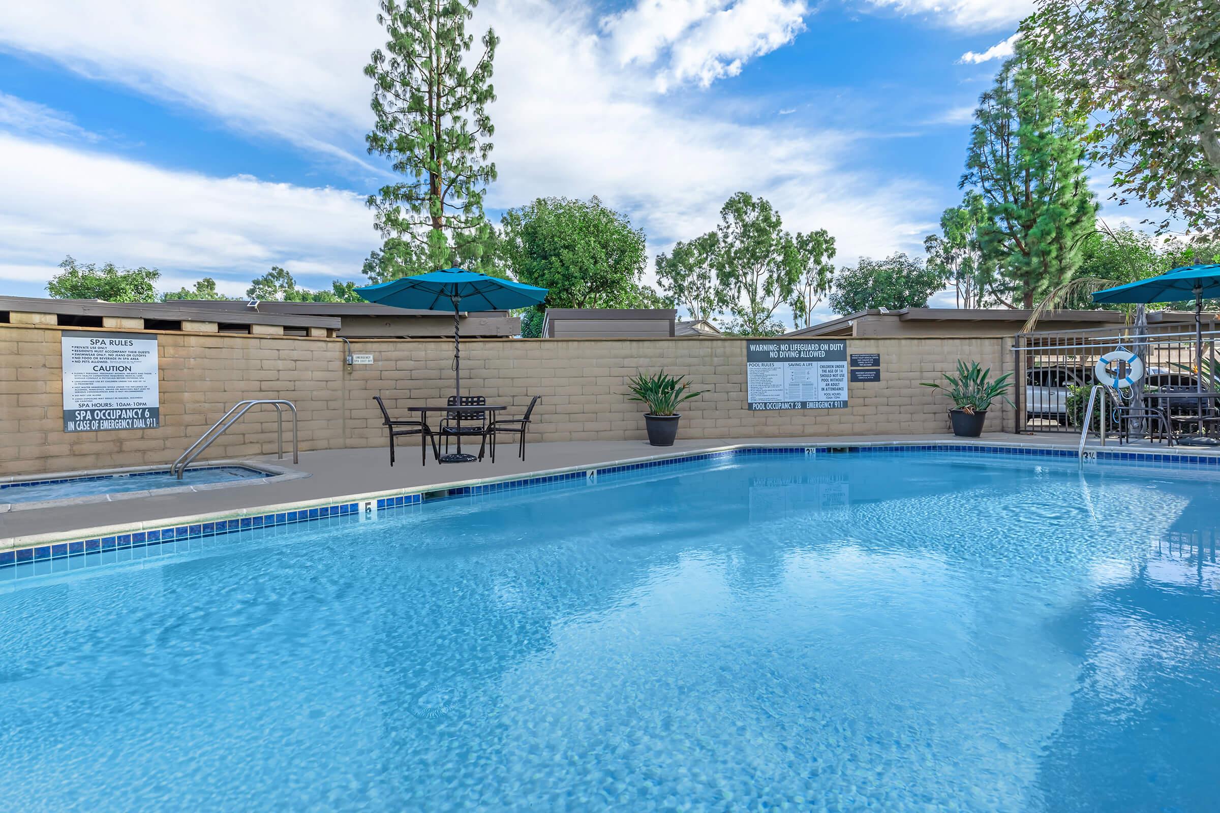 A clear blue swimming pool surrounded by lounge chairs, tables with umbrellas, and lush greenery. Signs are posted on the walls near the pool, and a few trees are visible in the background under a partly cloudy sky. The serene setting invites relaxation and leisure activities.