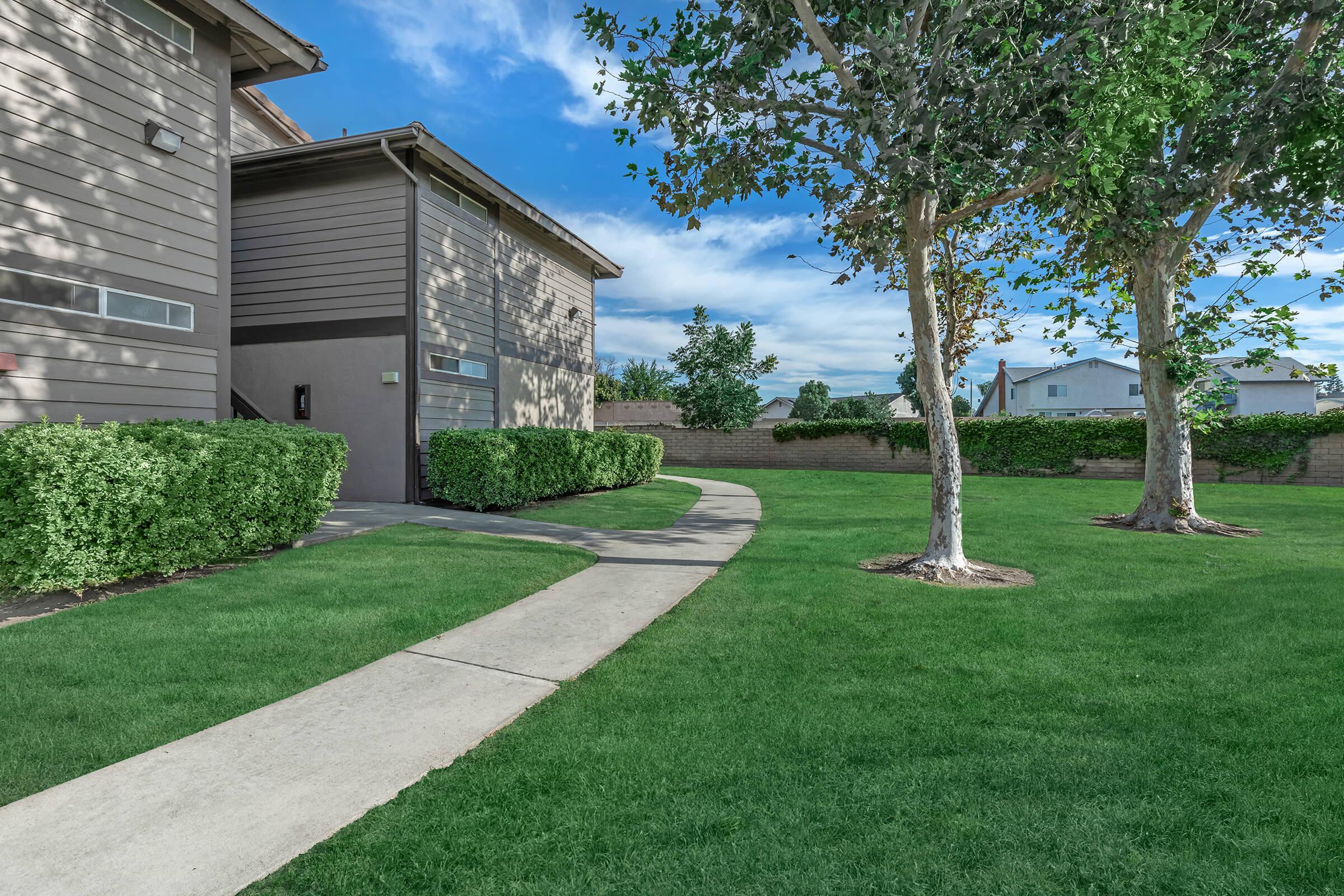 A well-maintained outdoor area featuring a curved concrete walkway leading to a gray-brown building. Lush green grass surrounds the path, with neatly trimmed hedges and two trees providing shade. In the background, additional buildings are visible under a clear blue sky with some clouds.
