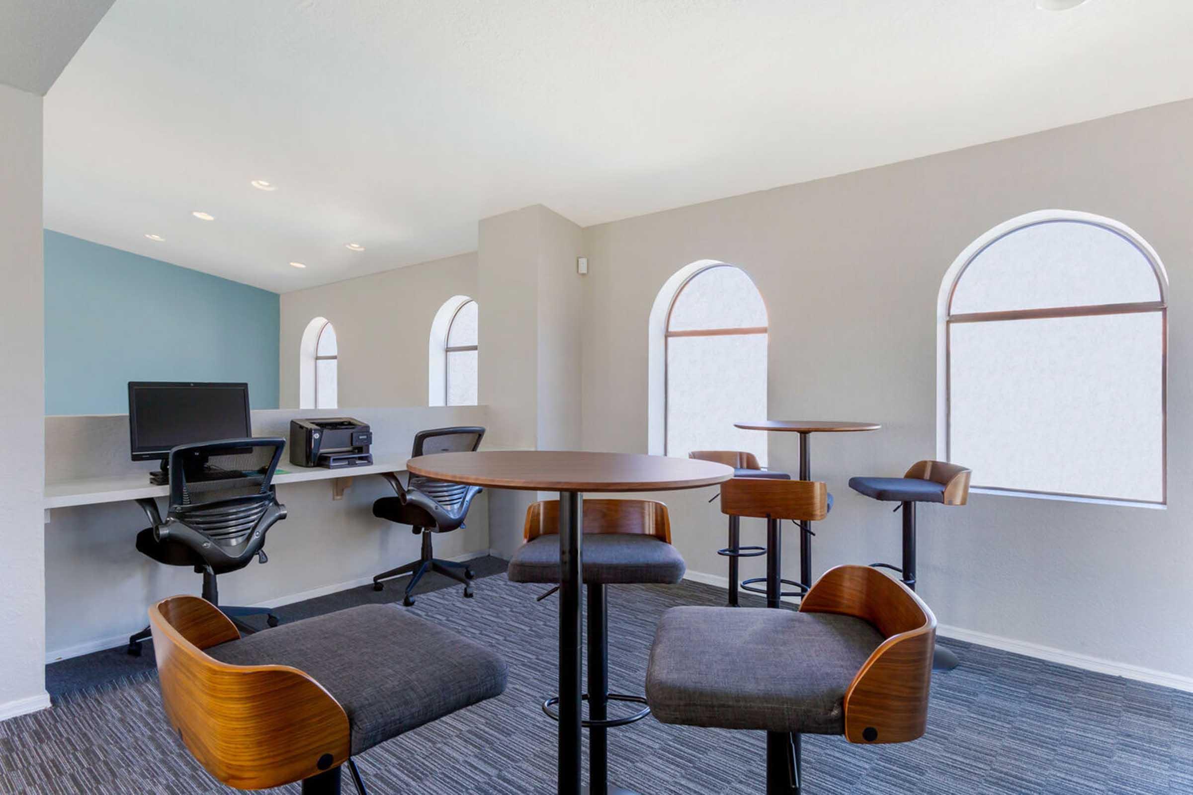 Modern workspace featuring high tables and chairs, two computer stations with monitors, and large arched windows allowing natural light.