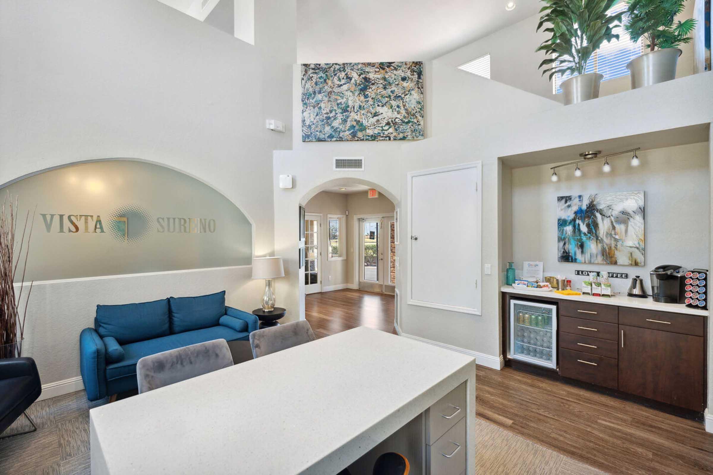 Interior view of a modern lobby space featuring a blue couch, a stylish coffee bar with a refrigerator, and decorative artwork on the walls.