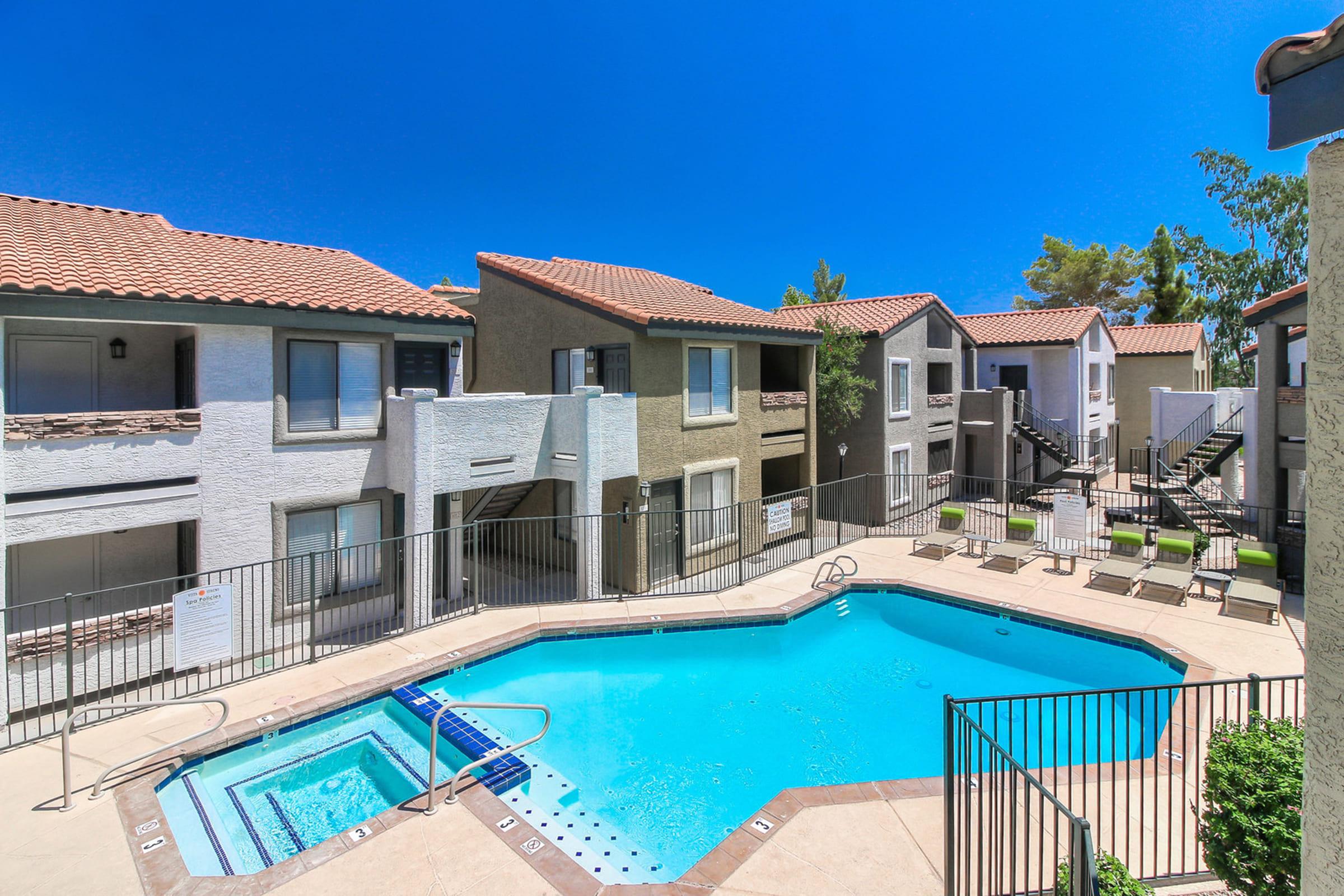 A view of a residential complex featuring a blue swimming pool surrounded by tan lounge chairs. The pool area is enclosed by a black fence, and several multi-story apartment buildings with red tile roofs are visible in the background under a clear blue sky.