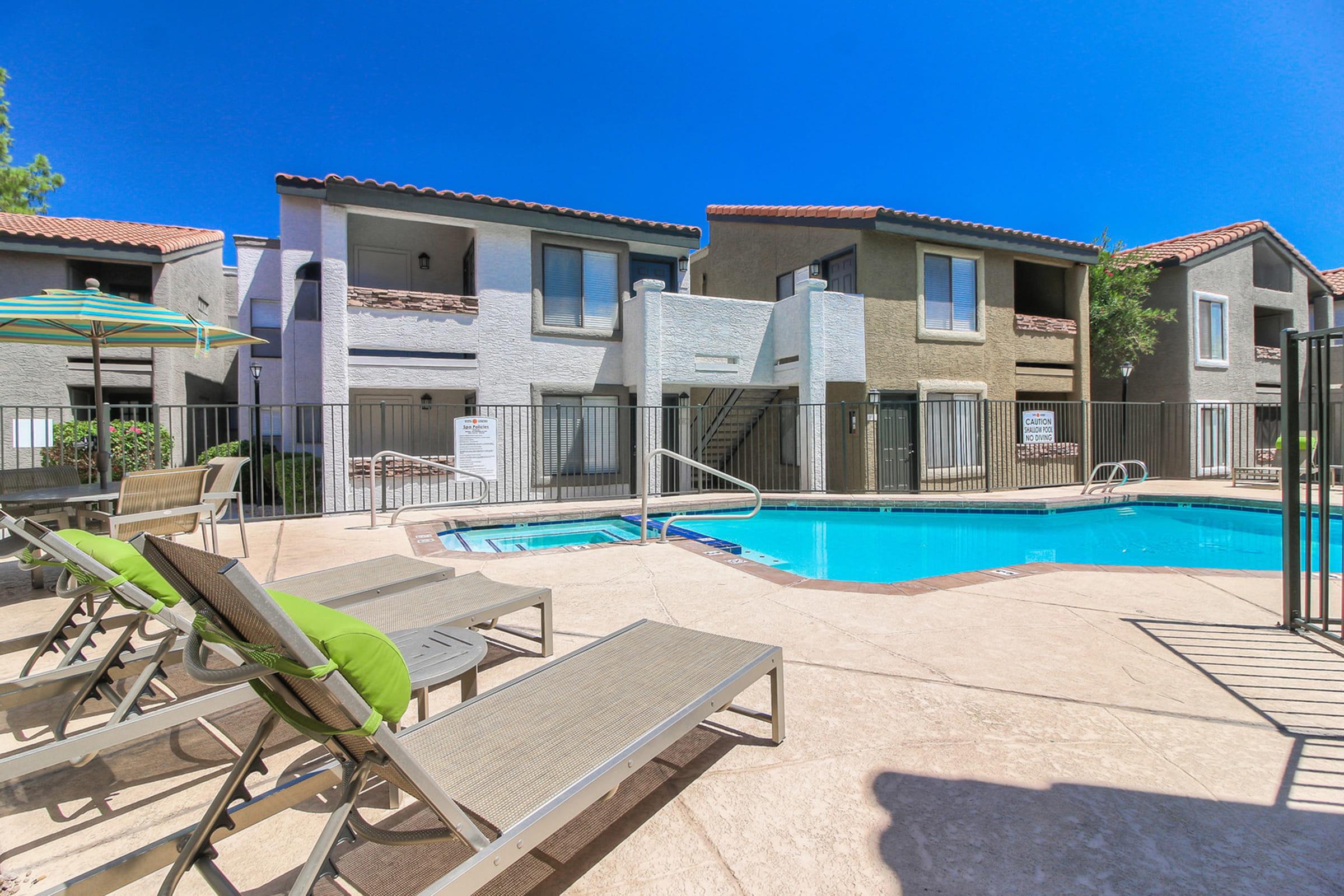 A swimming pool surrounded by lounge chairs and green umbrellas, with two-story apartment buildings in the background.