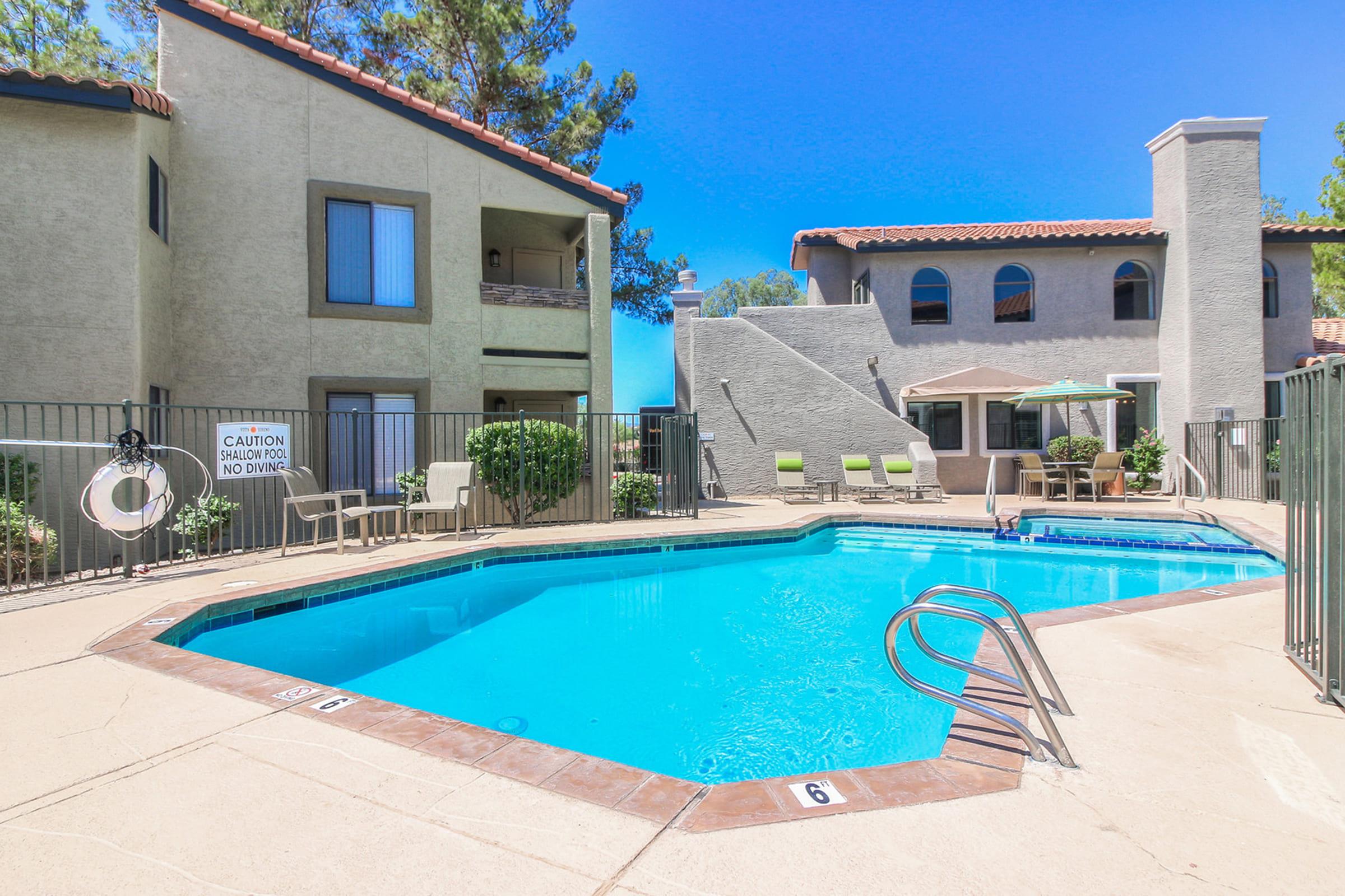 A sunny outdoor pool area at an apartment complex, featuring a clear blue swimming pool with a ladder, lounge chairs around the pool, and a building with balconies in the background. Lush greenery and a caution sign are also visible.
