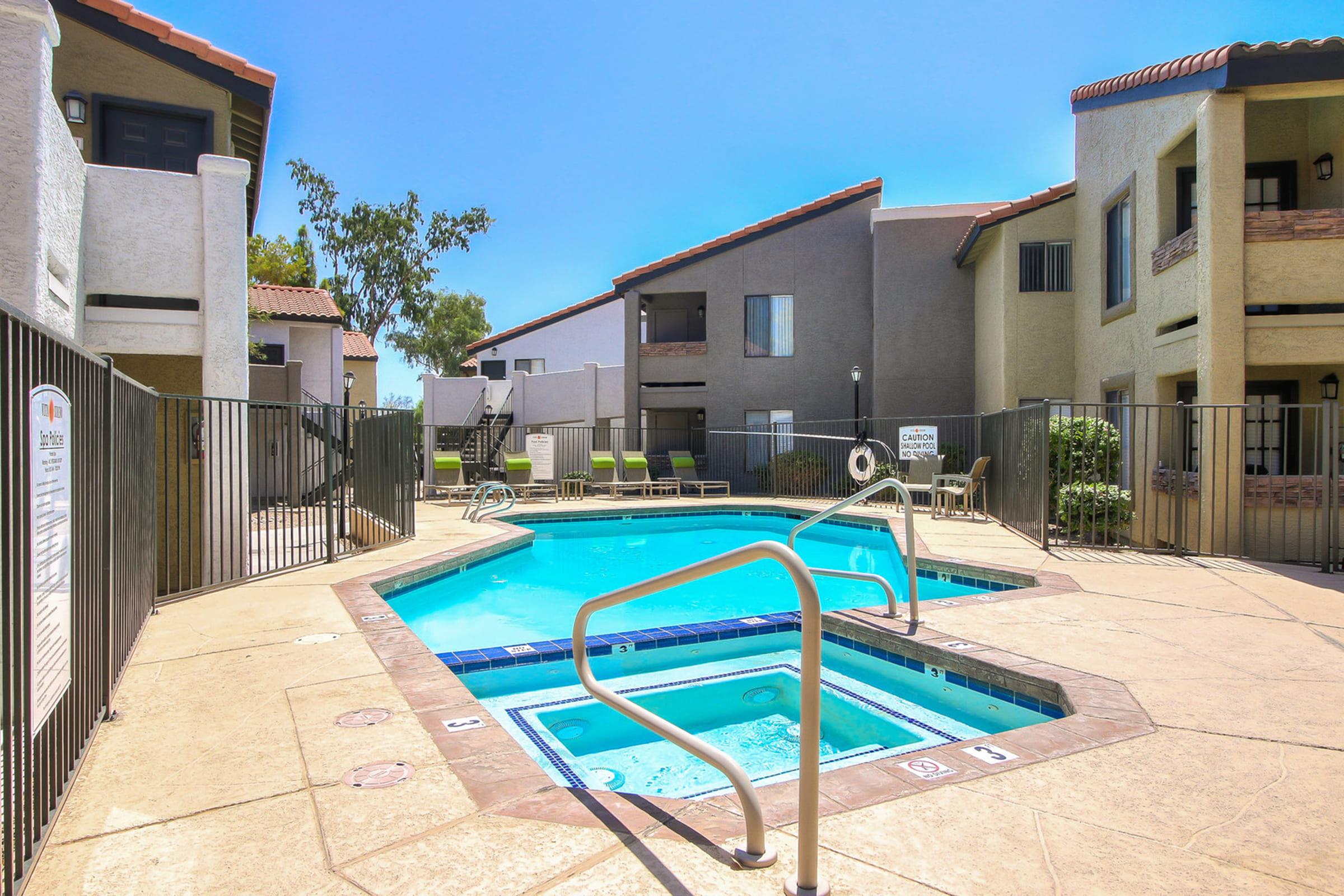 A clear, inviting pool area surrounded by a gated enclosure, featuring a hot tub and lounge chairs.