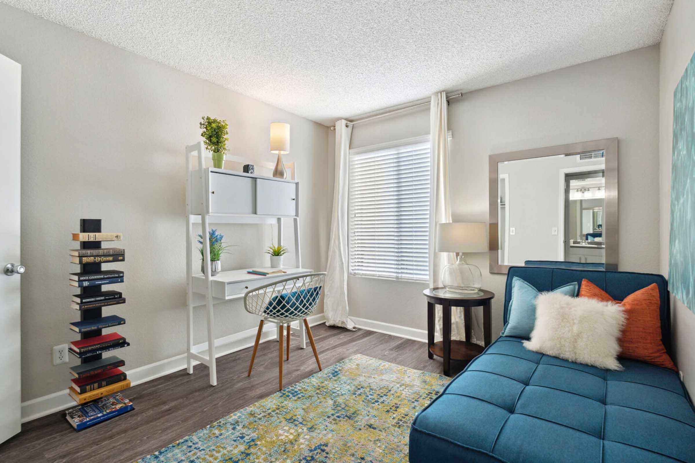 A cozy bedroom featuring a blue chaise lounge with decorative pillows, a modern desk with a chair, a lamp, and a tall stack of books.