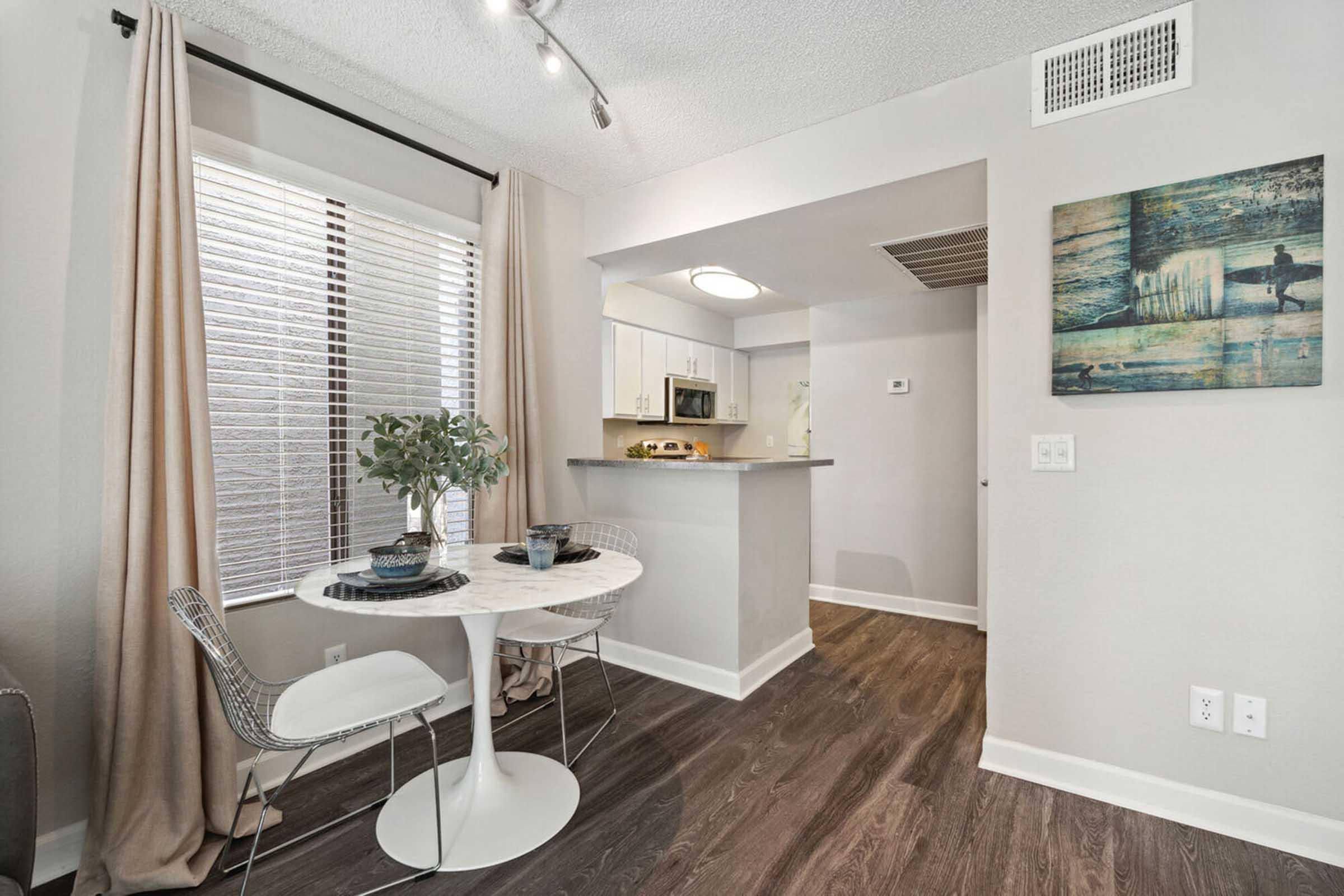 A modern dining nook featuring a round white table with two stylish chairs. Natural light streams through large windows with blinds, complemented by light gray walls.