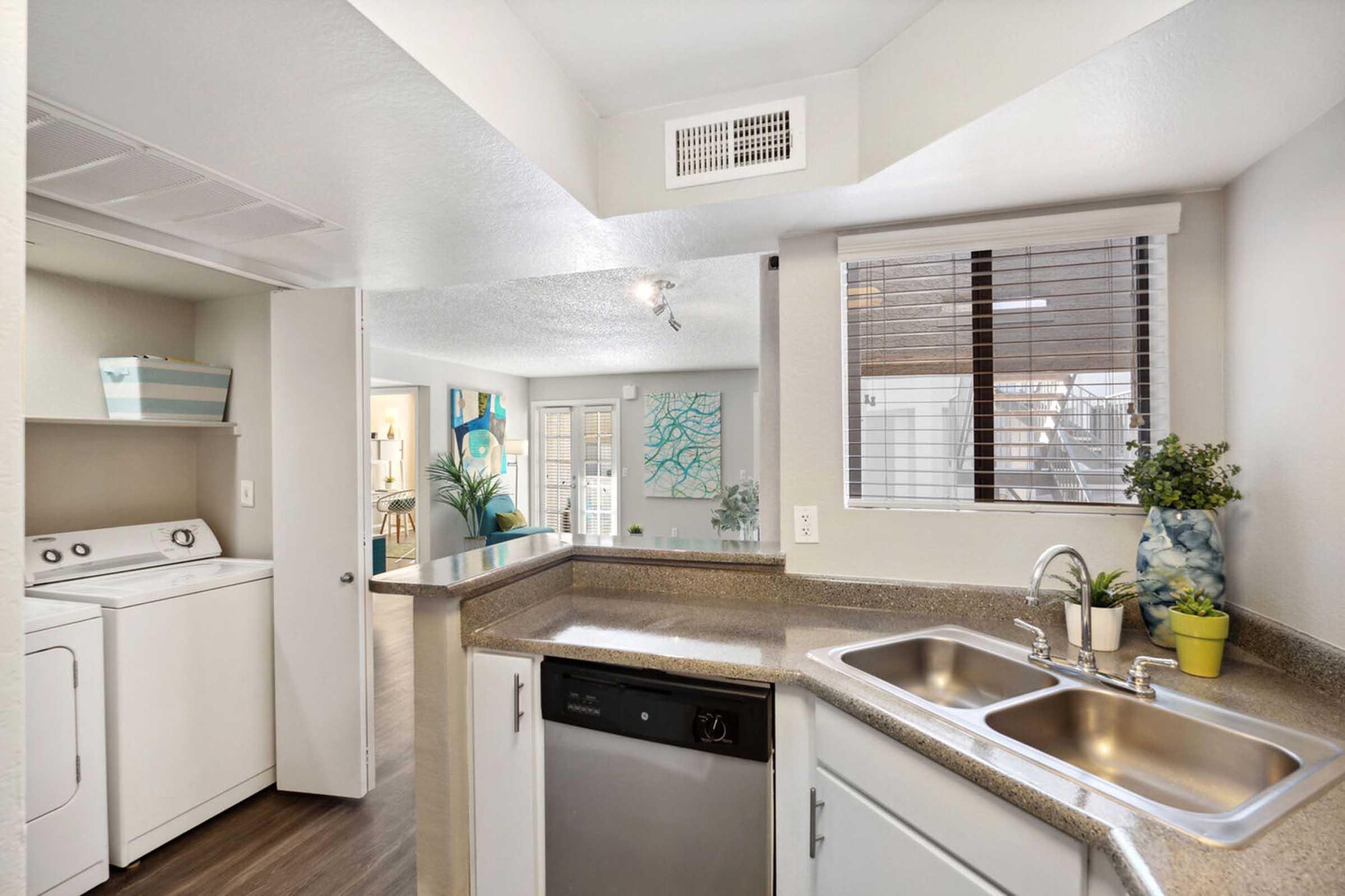 A modern kitchen featuring a double sink, dishwasher, and laundry area. The room has light-colored walls, wooden flooring, and a large window with blinds.