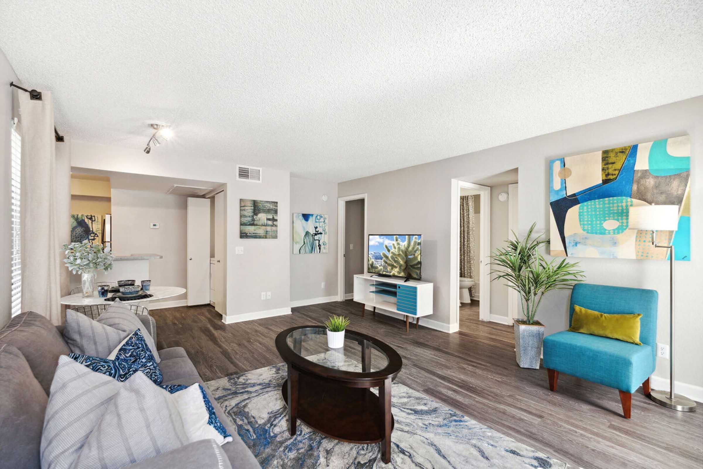 A modern living room with a gray sofa adorned with blue cushions. There’s a round coffee table in front, and a flat-screen TV on a white console.