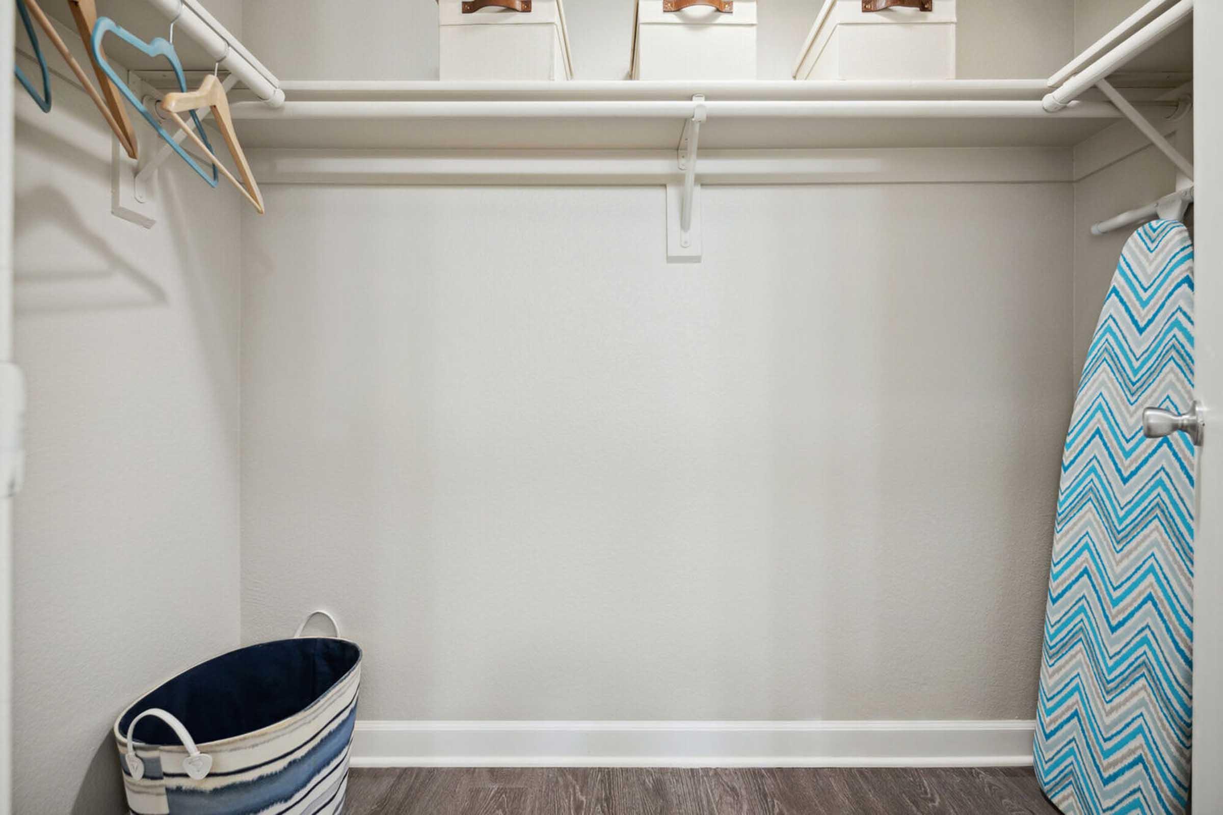 A clean and organized closet featuring a metal rod with wooden hangers, an empty space, a blue and white striped laundry basket, and an ironing board with a chevron pattern.