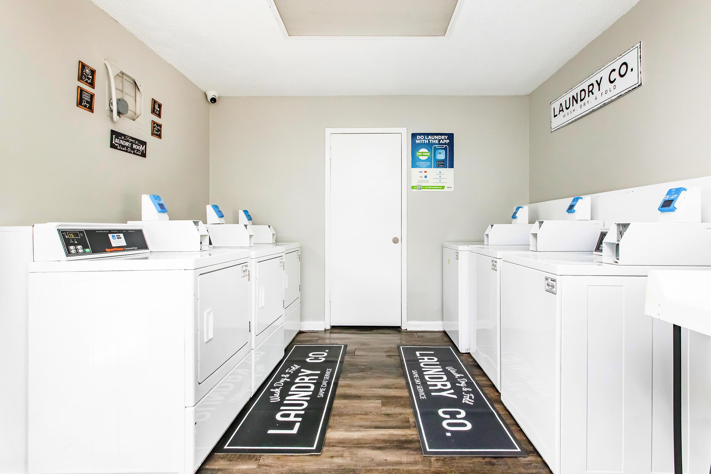 A bright and clean laundry room featuring several white washing machines lined up against the walls. The space includes two mats with "LAUNDRY CO." printed on them, light gray walls, and a door at the end of the room. Signs on the walls provide information about laundry services.