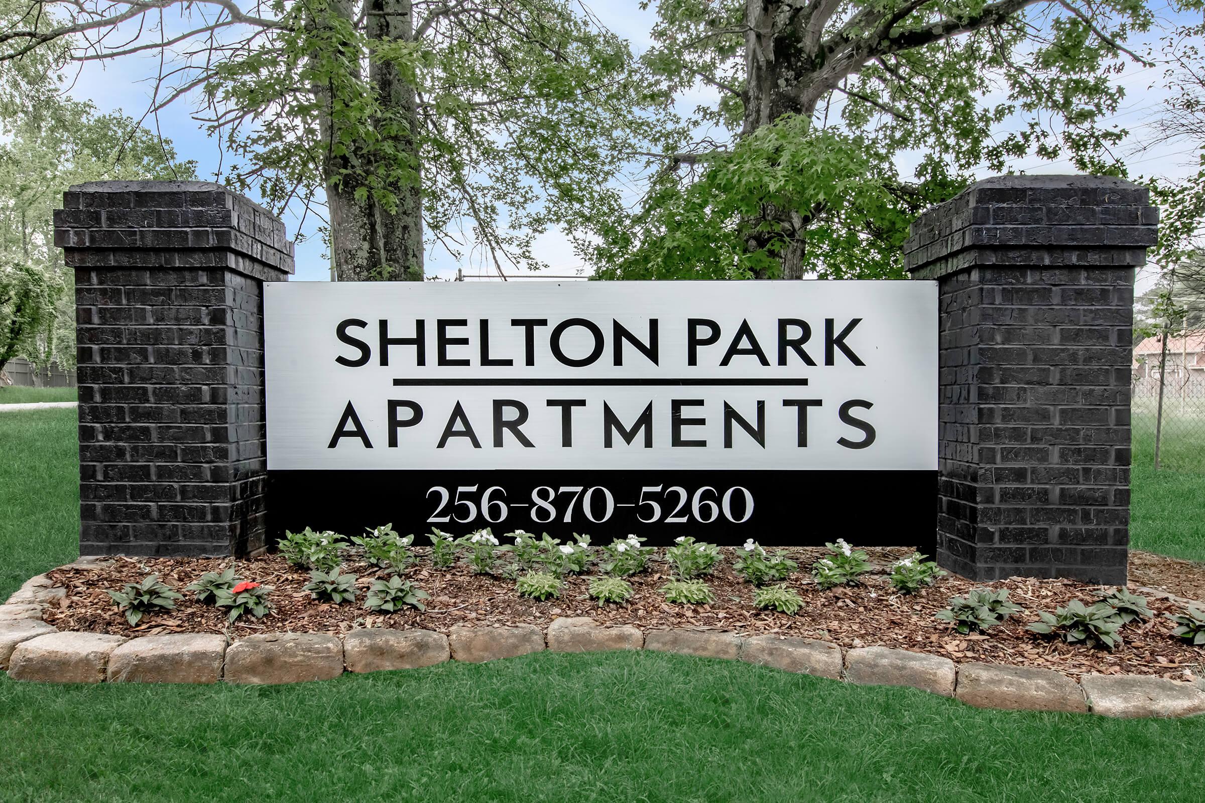 Sign for Shelton Park Apartments showcasing the name prominently. The sign features black stone pillars on either side and decorative plants in the foreground. Contact number displayed: 256-870-5260. Surrounding area includes greenery and trees.