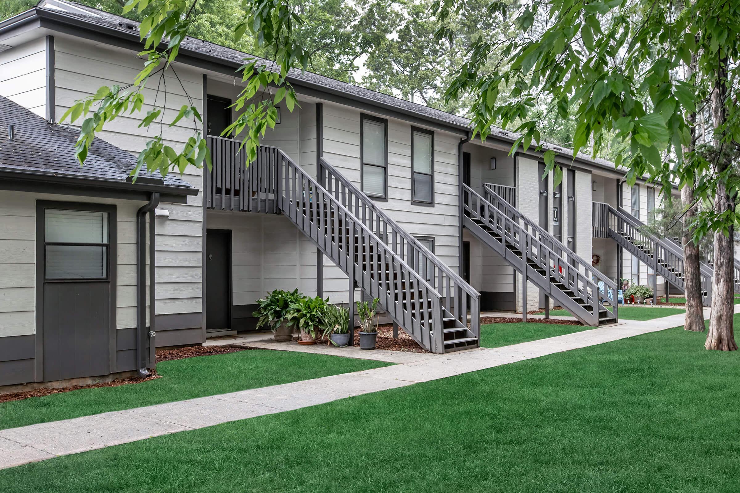 A view of a residential building with two stories, featuring gray exterior walls and wooden staircases leading to the upper level. The landscape includes well-manicured green grass and potted plants along the walkway, which connects the entrances of the apartments. Trees provide shade in the surroundings.