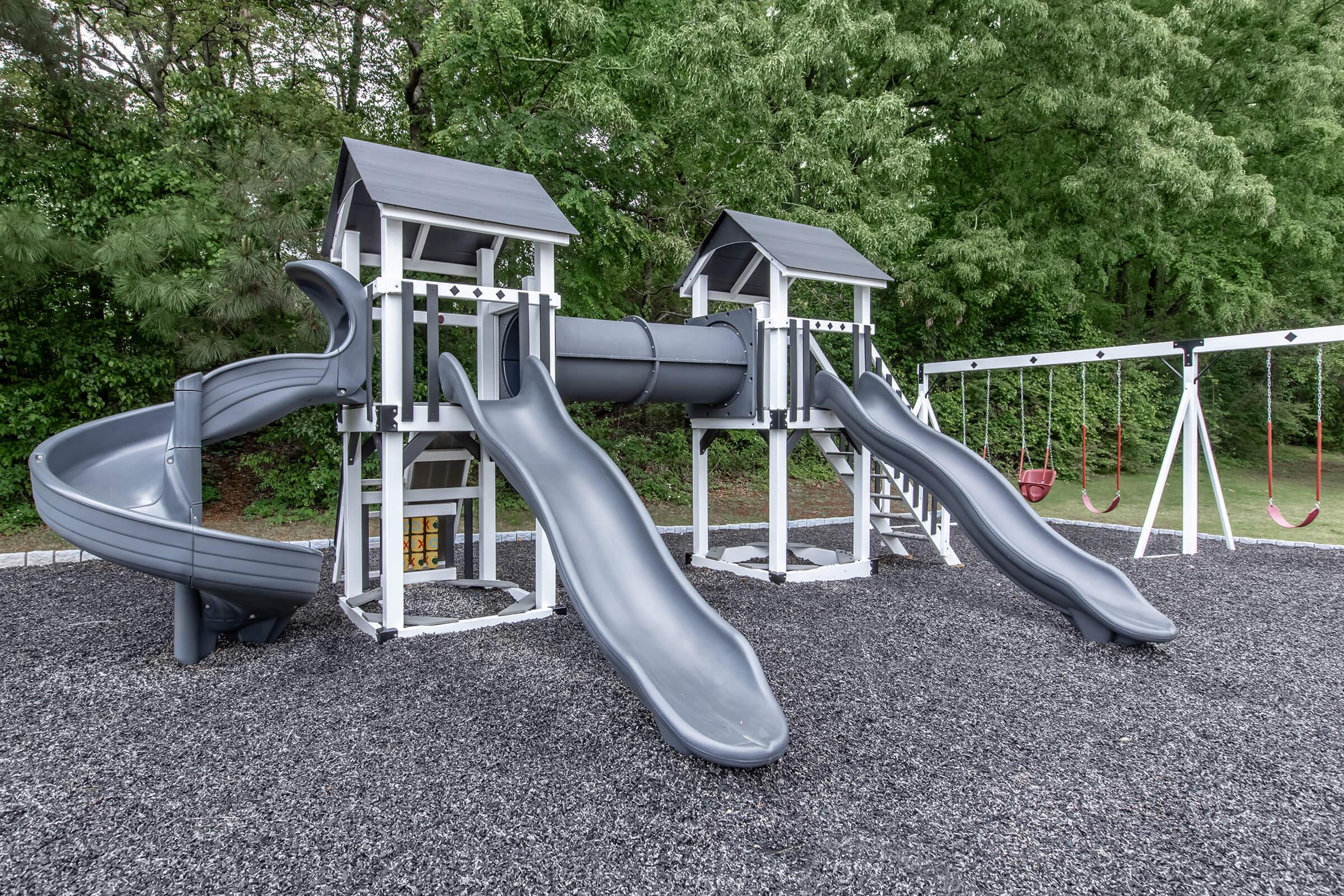A playground featuring two gray slides, a central tunnel, and a swing set. The structure is set on a gravel surface with green trees in the background, creating a vibrant outdoor play area. Suitable for children to enjoy various activities.