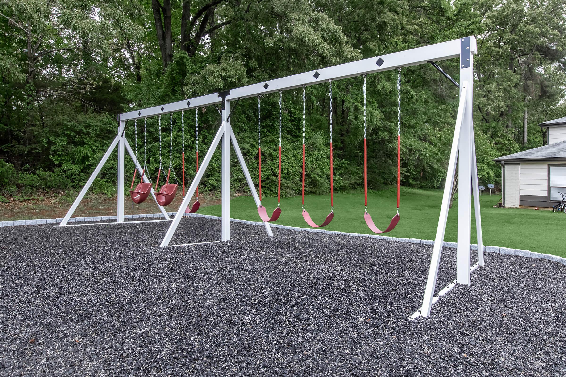 A playground featuring a set of red swings hanging from a white frame, situated on a gravel surface. Lush green trees are visible in the background, and a building can be seen to the right. The scene is tranquil, ideal for outdoor play.