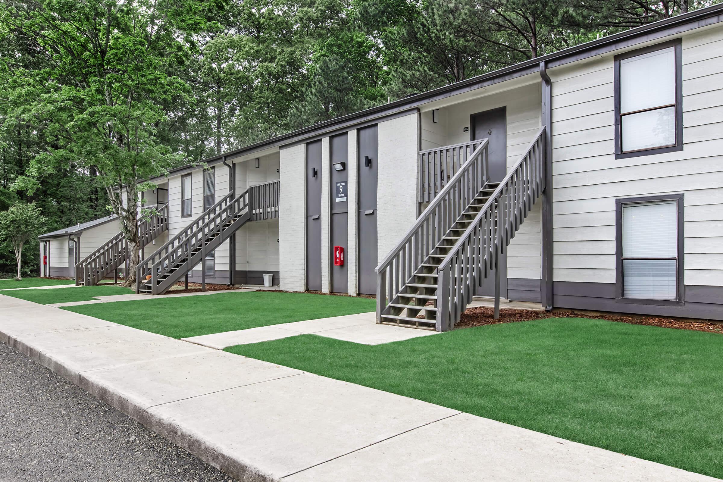 A view of a residential building with two sets of stairs leading to upper-level apartments. Lush green grass surrounds the concrete pathway and the exterior features a modern design with a light color palette. Trees are visible in the background, providing a natural setting.