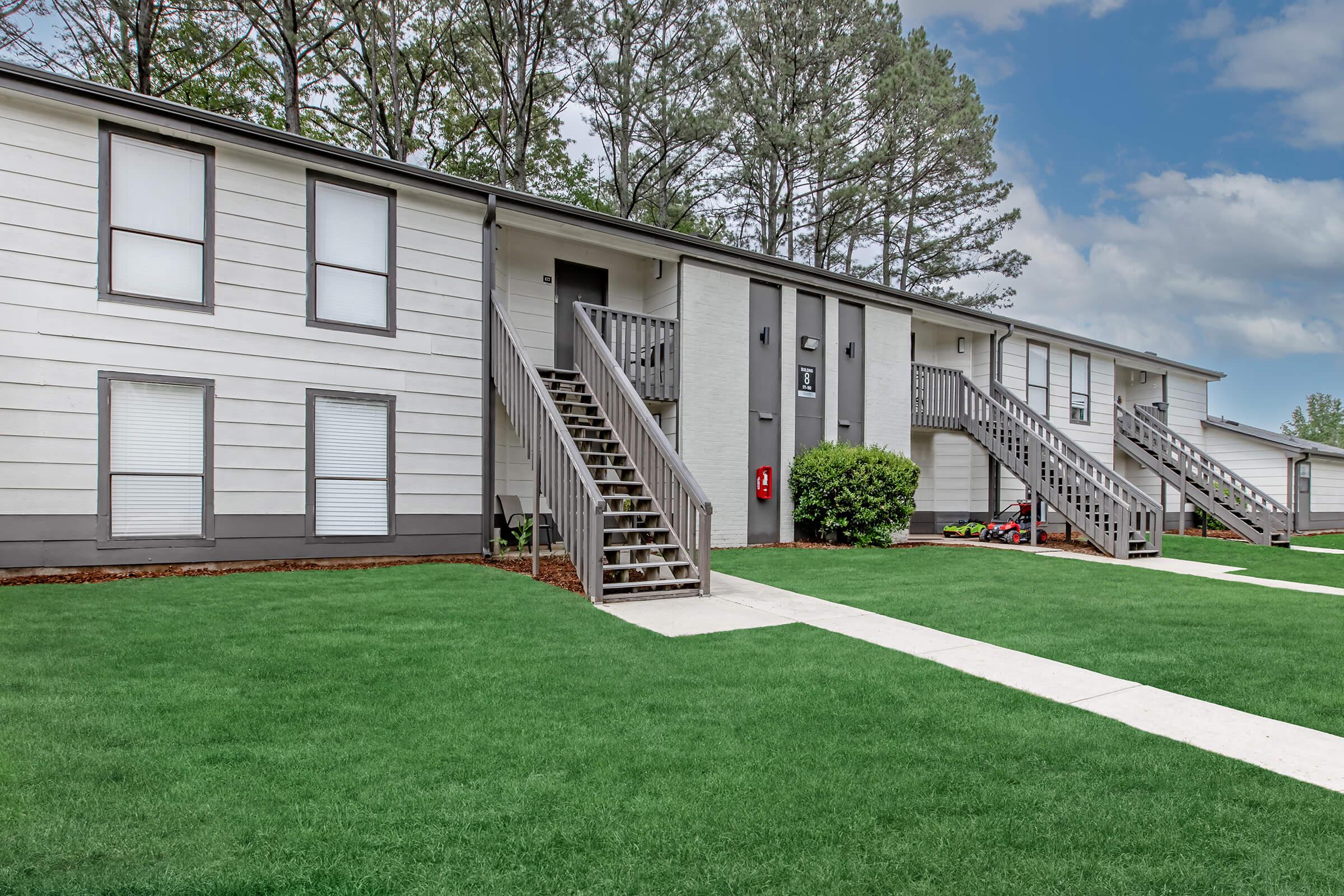 A view of a two-story apartment building with a grassy lawn. The building features a series of stairs leading to individual units, with windows and landscaped areas in front. The sky is slightly cloudy, and the surrounding trees add to the greenery of the scene.