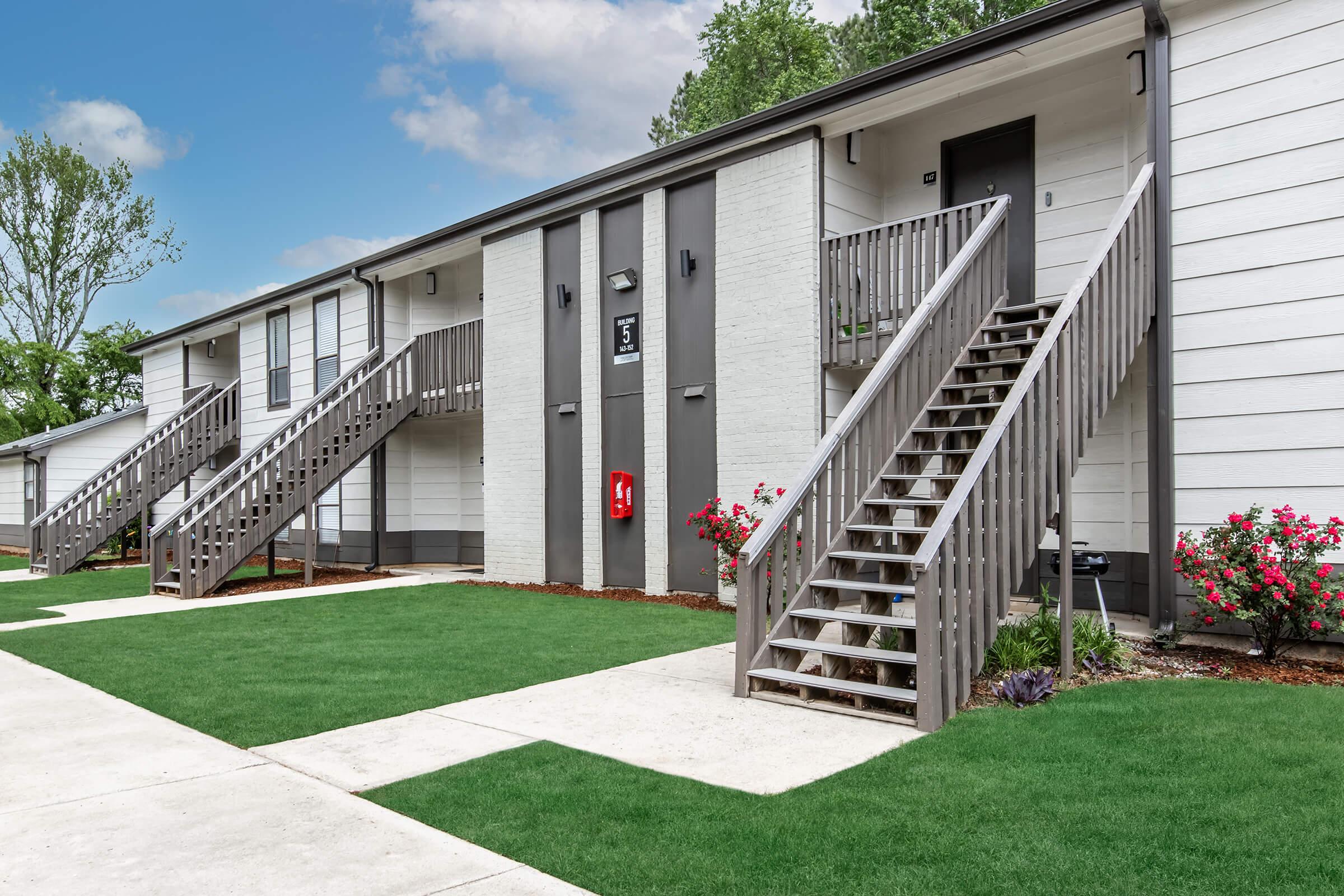Two-story apartment building with a modern design featuring gray and white exteriors. There are wooden staircases leading to the upper units, flanked by landscaped green lawns and flower beds. The setting is well-maintained, with a clear sky above.