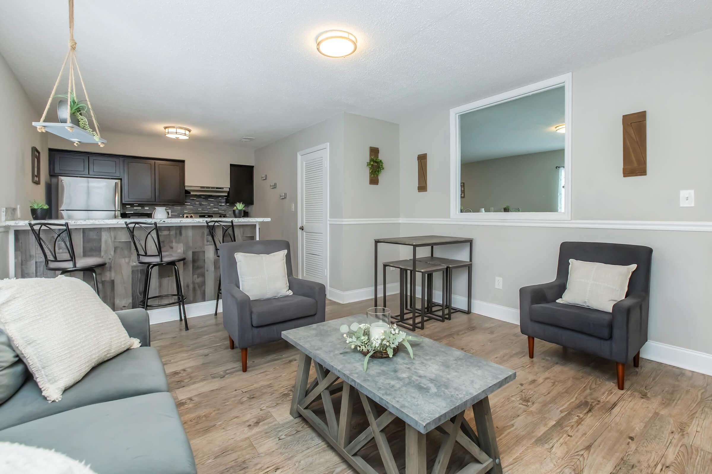 A modern living room featuring two dark gray armchairs and a coffee table with a stone top. Behind, there is a small dining area with bar stools and a kitchen visible. The walls are light gray with decorative plants and framed artwork. The floor is a light wood finish, creating a cozy atmosphere.