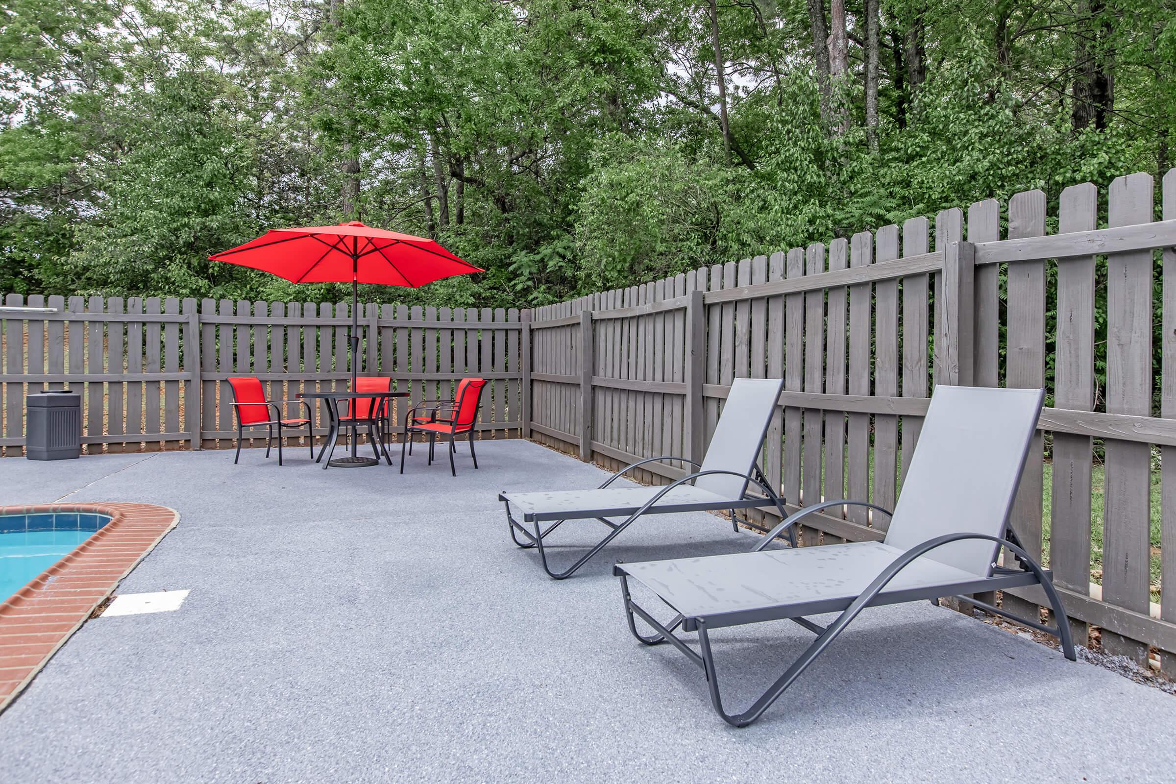 A poolside area featuring two gray lounge chairs, a round table with four red chairs, and a bright red umbrella. The space is enclosed by a wooden fence and surrounded by lush greenery, creating a relaxing outdoor setting.