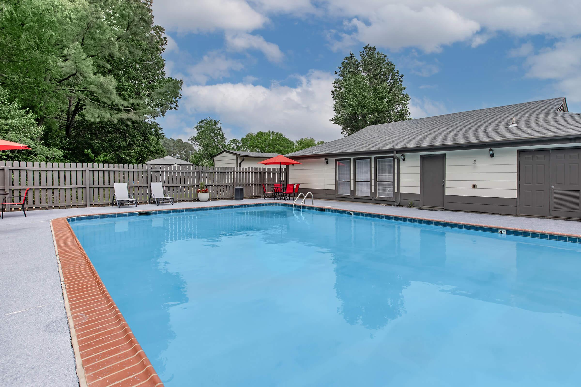 A clear blue swimming pool surrounded by a patio area. Lounge chairs are arranged beside the pool, and red umbrellas provide shade. In the background, a gray house with a porch and a wooden fence is visible, along with trees adding greenery to the scene. The sky is partly cloudy.