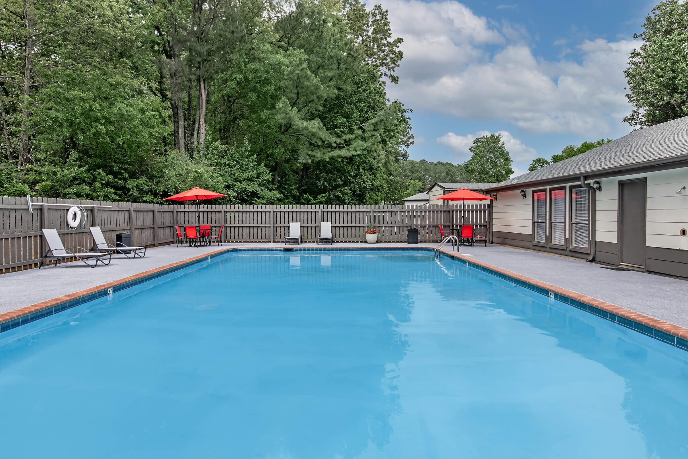 A clear blue swimming pool surrounded by a gray patio. Lounge chairs are positioned beside the pool, and two red umbrellas provide shade. In the background, a wooden fence and trees are visible, along with a building that has several windows. The sky is partly cloudy.