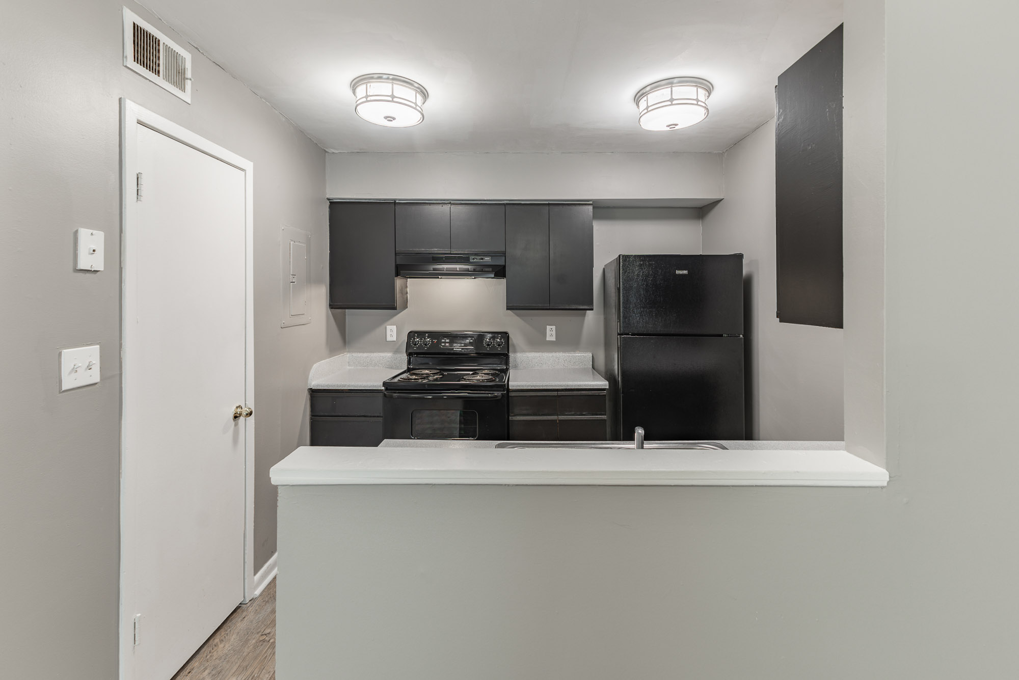 A modern kitchen featuring dark cabinets, a black stove, and a black refrigerator. The kitchen has light gray walls and recessed lighting. A small countertop separates the kitchen from an adjacent room, with a door leading to another space.