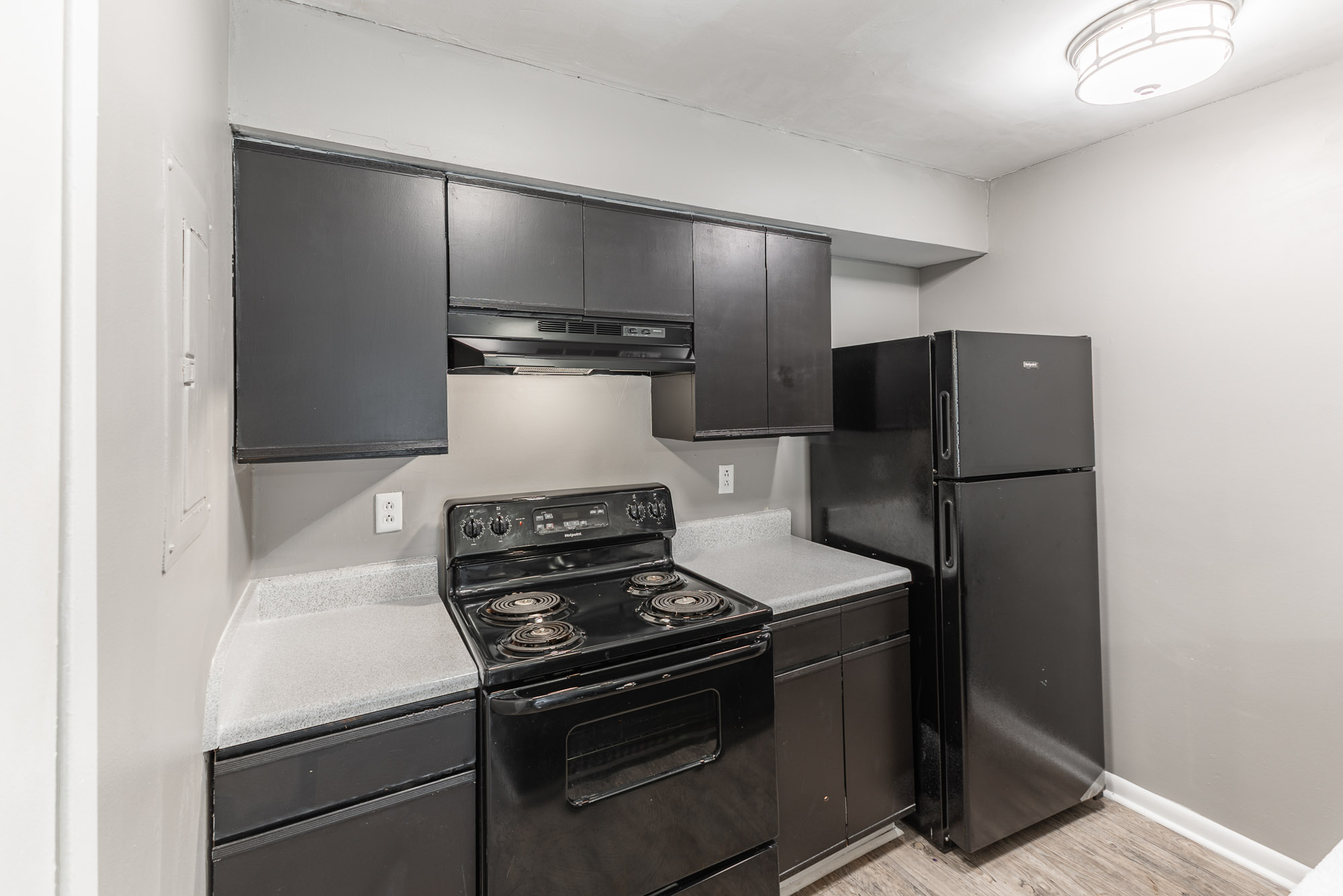 A modern kitchen featuring black cabinets, a black stove and oven, a black microwave, and a black refrigerator. The countertops are light gray, and the walls are painted a neutral color, creating a contemporary look. A ceiling light fixture adds illumination to the space.