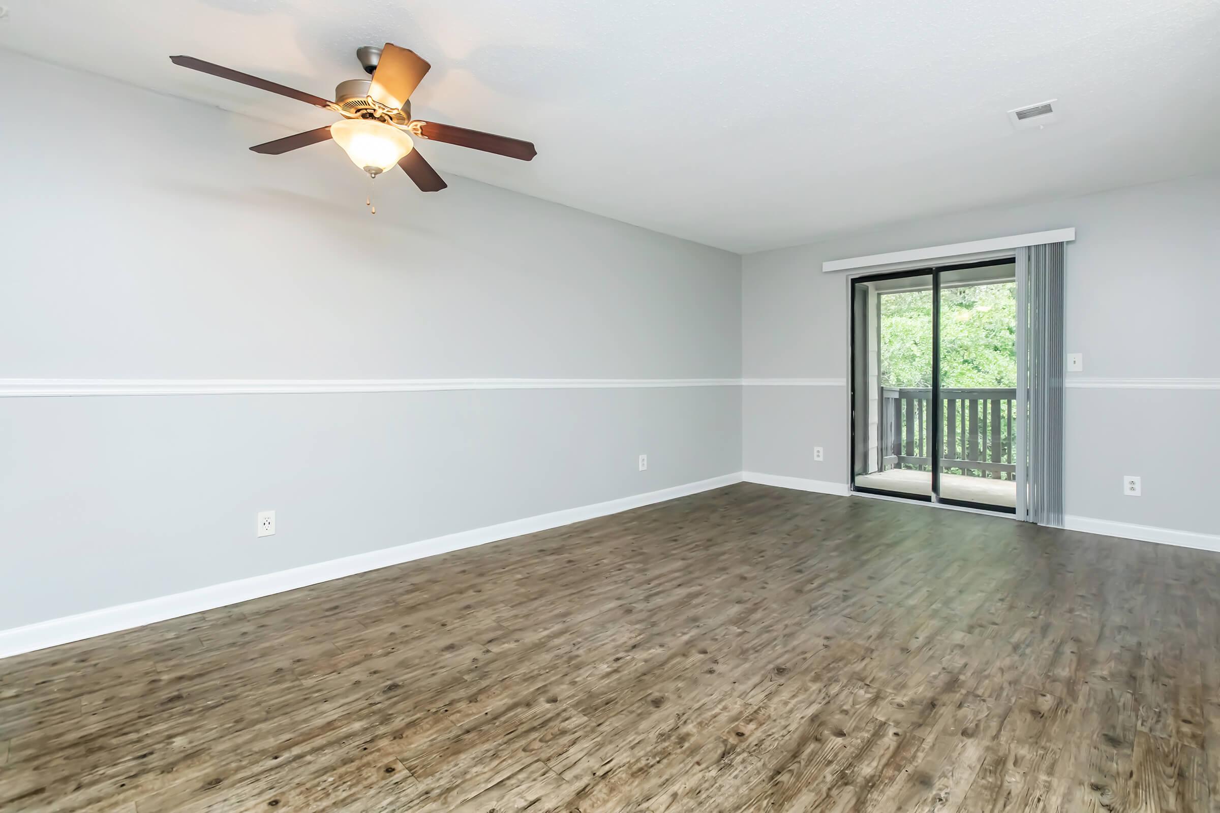Spacious living room featuring a ceiling fan, light-colored walls, and a balcony with sliding glass doors. The floor is made of wood-like laminate, and the room is well-lit with natural light from the outdoor view.