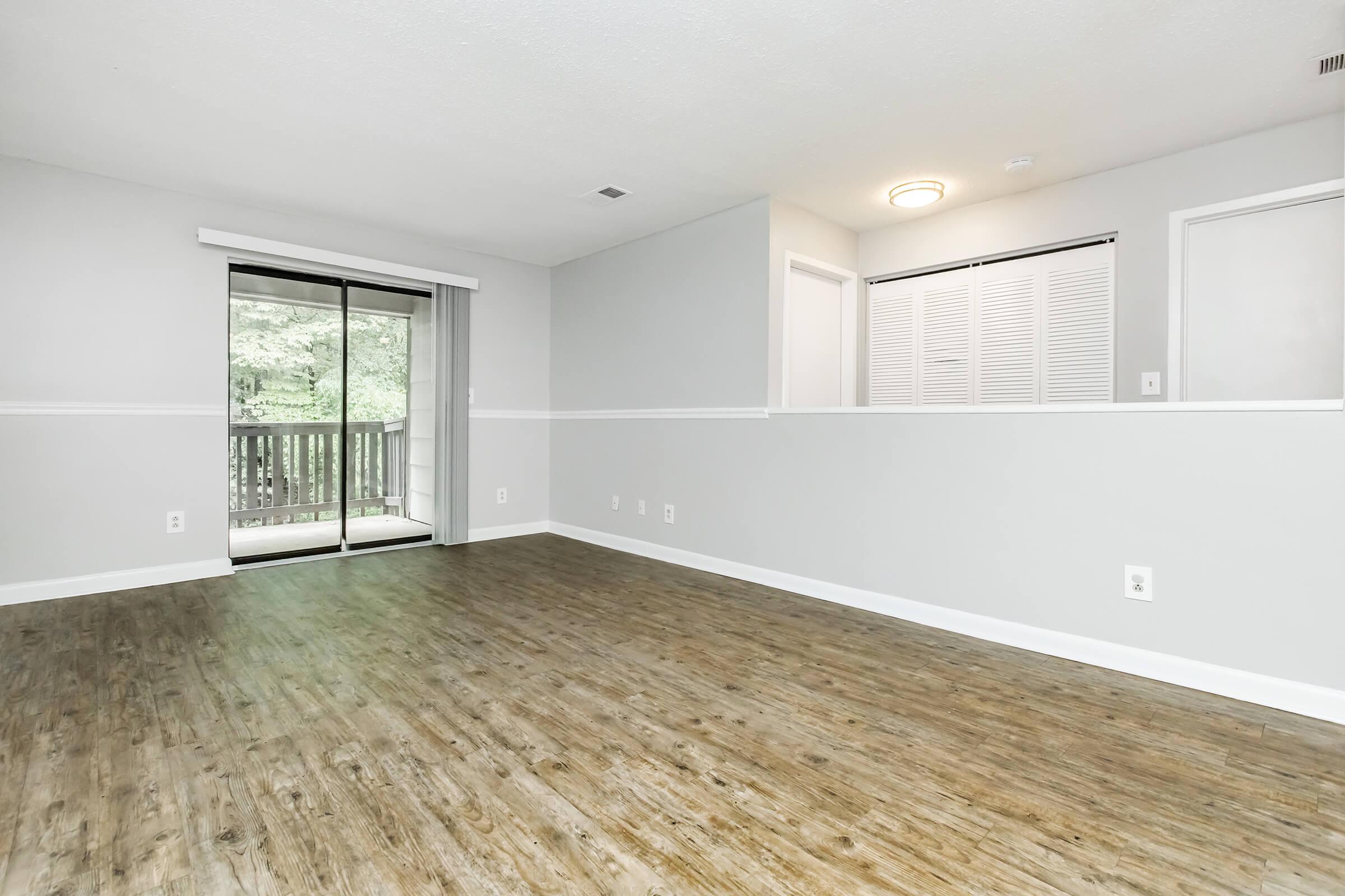 A spacious, empty living room featuring light gray walls, wooden flooring, and large sliding glass doors leading to an outdoor balcony. The room has minimalist décor with a soft overhead light and a small utility area with closet doors. Natural light illuminates the space, creating an inviting atmosphere.