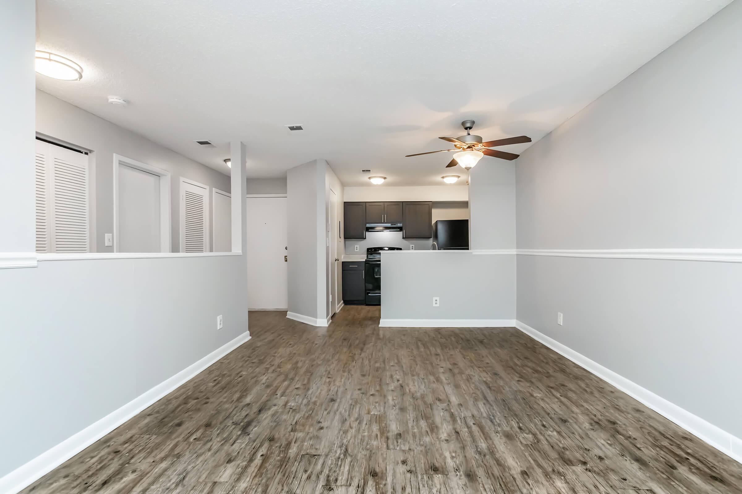 Spacious living area with hardwood-like flooring, featuring a ceiling fan. The room is painted in light grey tones and has white wainscoting. A kitchen area is visible in the background, equipped with modern appliances and dark cabinetry. The layout is open and bright, suggesting a welcoming atmosphere.