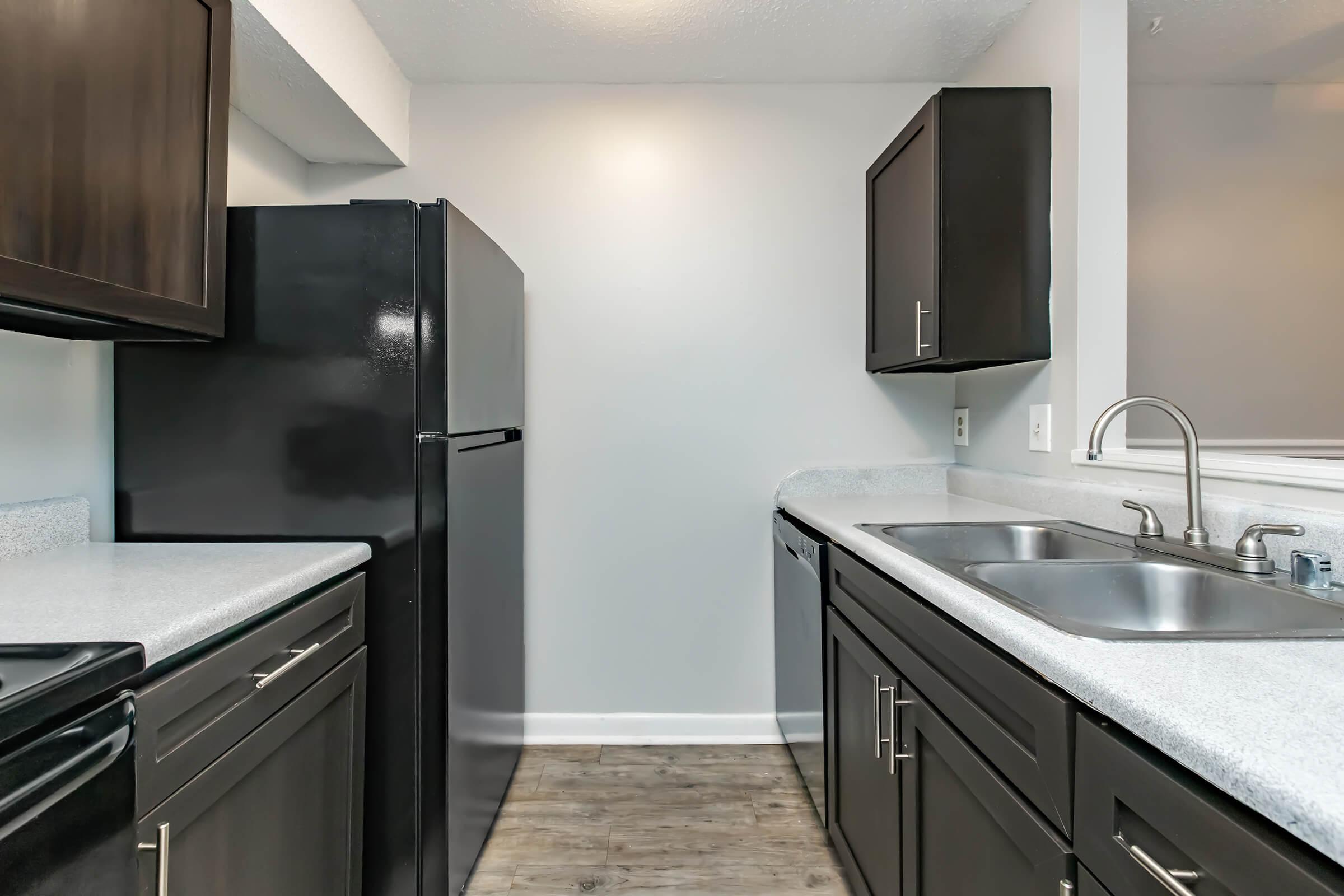 A modern kitchen featuring dark wood cabinets, a black refrigerator, and stainless steel double sinks. The countertops are light-colored, and the flooring is a wood-like finish. The space is well-lit with neutral wall colors, creating a sleek and contemporary atmosphere.