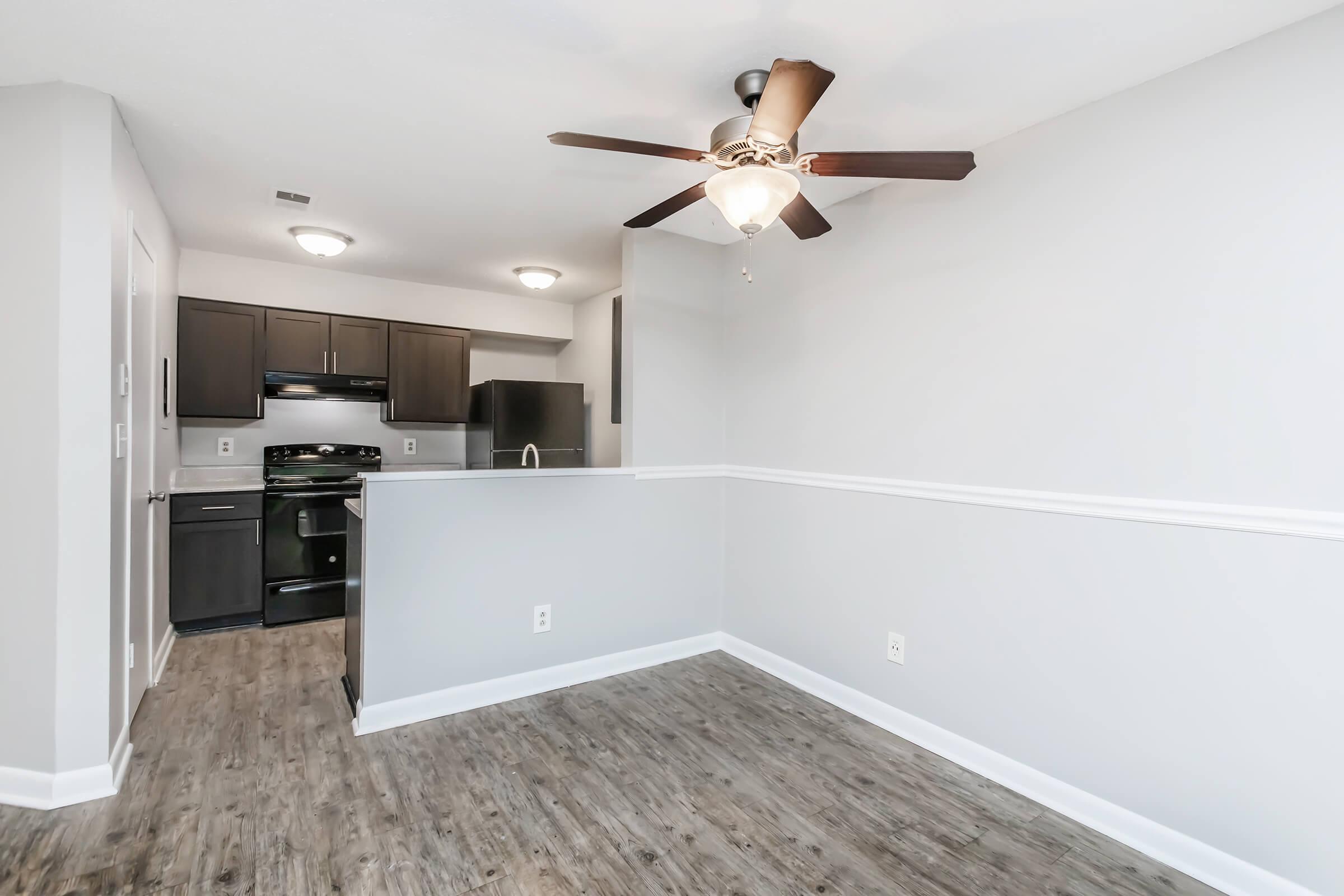 Interior of a modern apartment featuring a living area with wooden flooring, a ceiling fan, and an open kitchen with dark cabinetry and stainless steel appliances. The walls are painted light gray, and there is a decorative white molding accented along the edges.