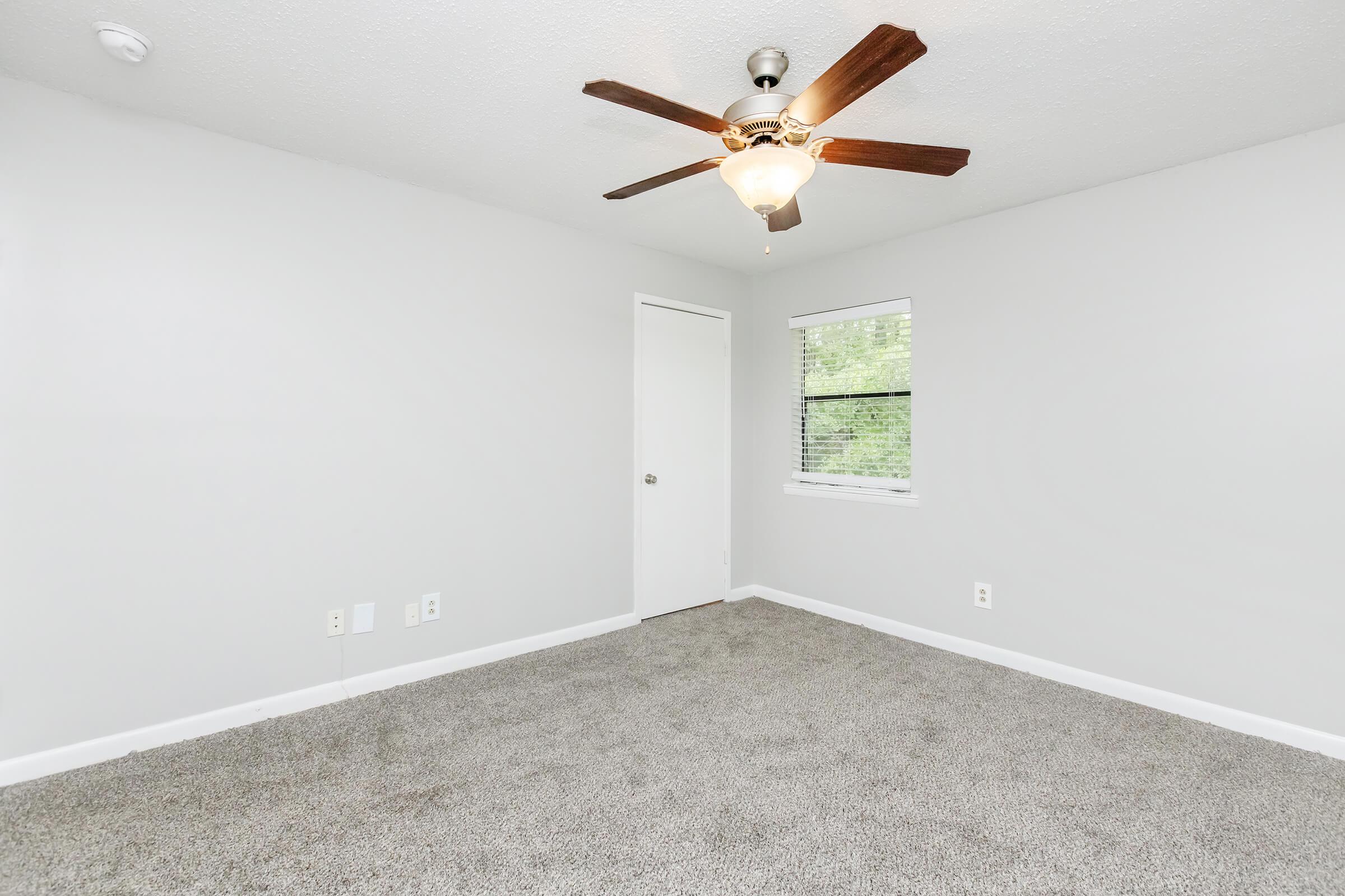 A vacant room with light gray walls and a soft carpeted floor. The ceiling features a ceiling fan with a light fixture. There is a closed white door on one side and a window with blinds letting in natural light. The space appears clean and ready for furnishings.