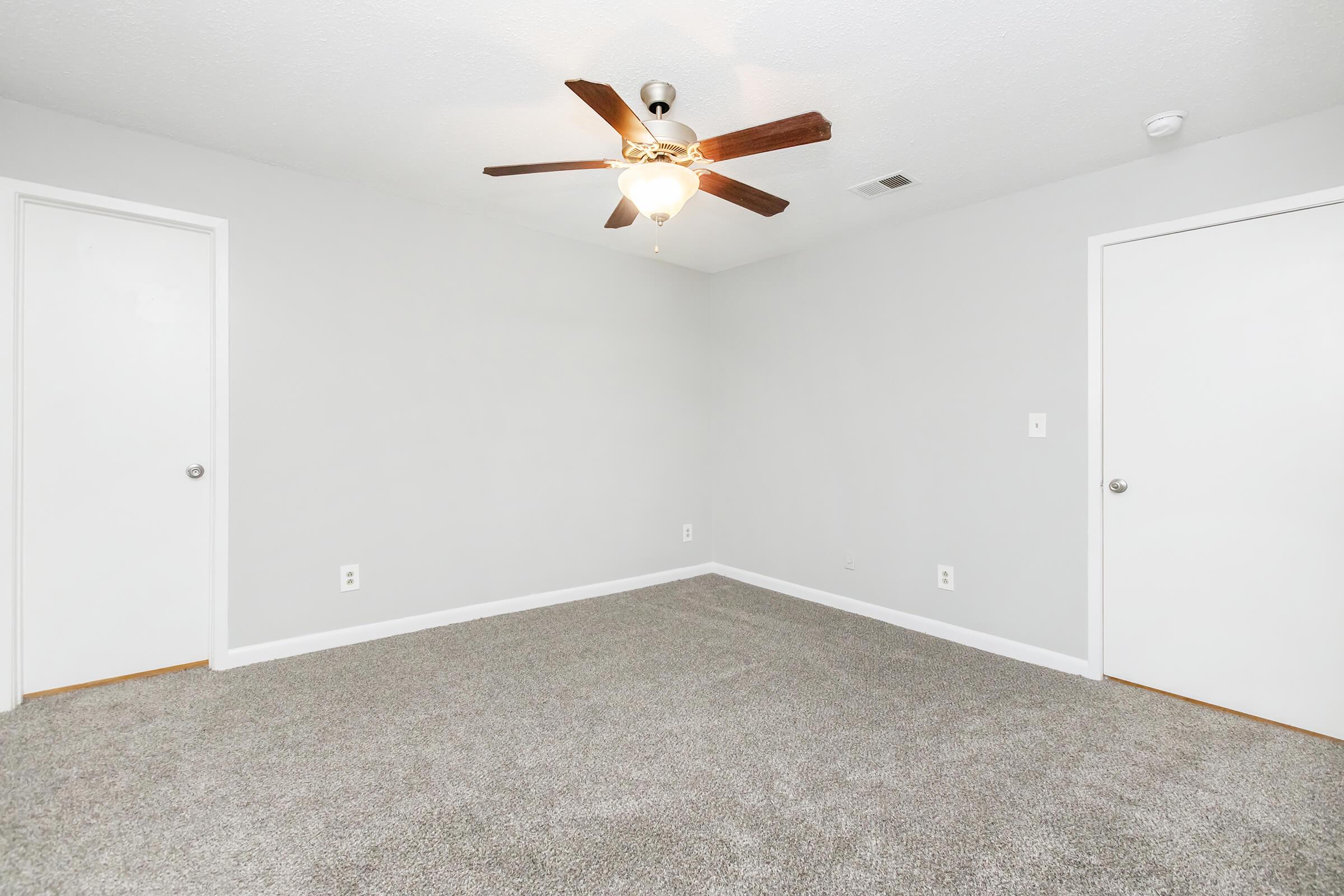A spacious, empty room with light gray walls and a carpeted floor. There is a ceiling fan with wooden blades in the center. Two white doors are visible on opposing walls, and the room is well-lit, giving it a bright and airy feel.