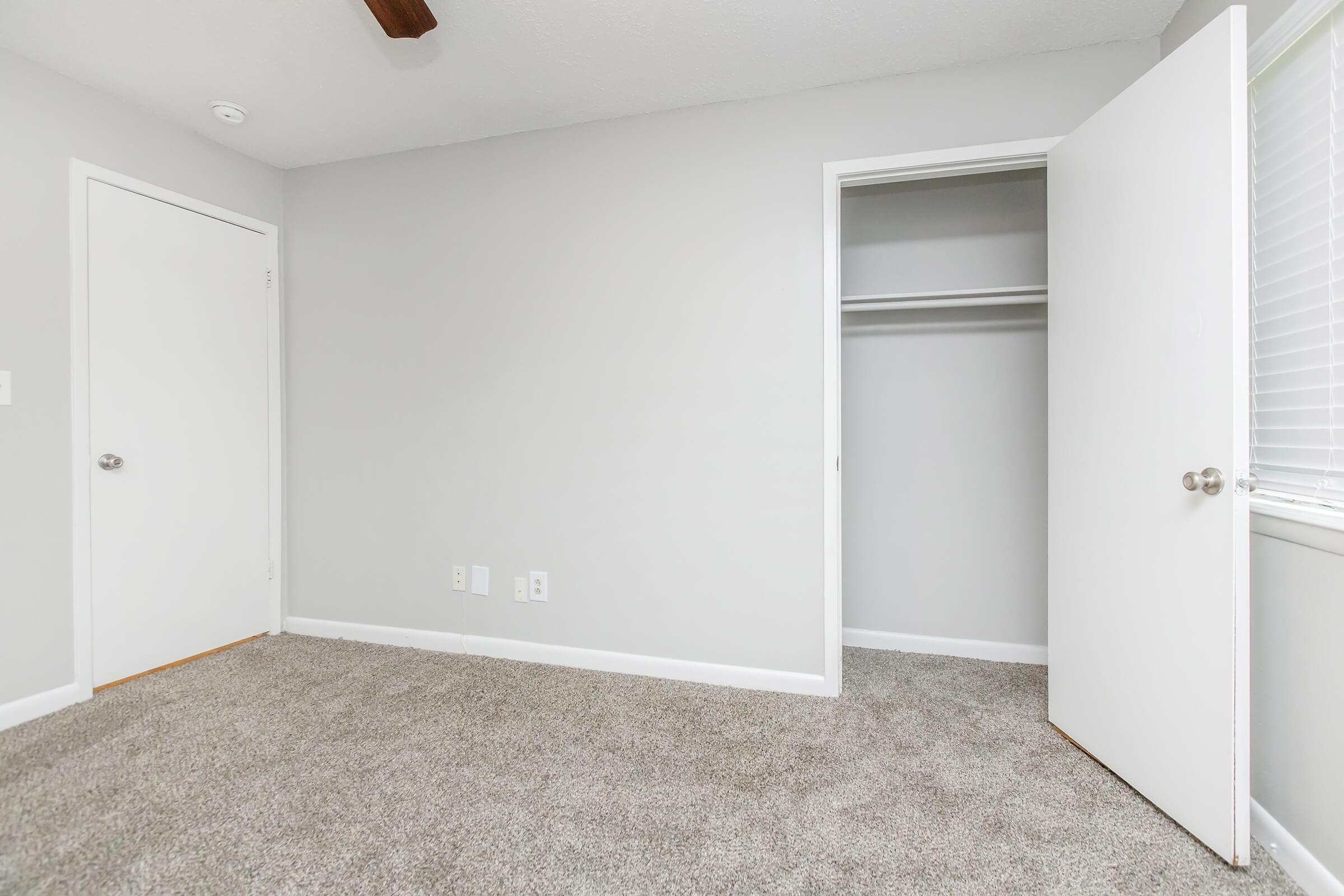 A well-lit, empty room with gray walls and beige carpet. There is a ceiling fan and a closed door on one side, while another door leads to a closet on the opposite side. Natural light enters through a window with white blinds. The space is clean and minimalist, ready for furniture.