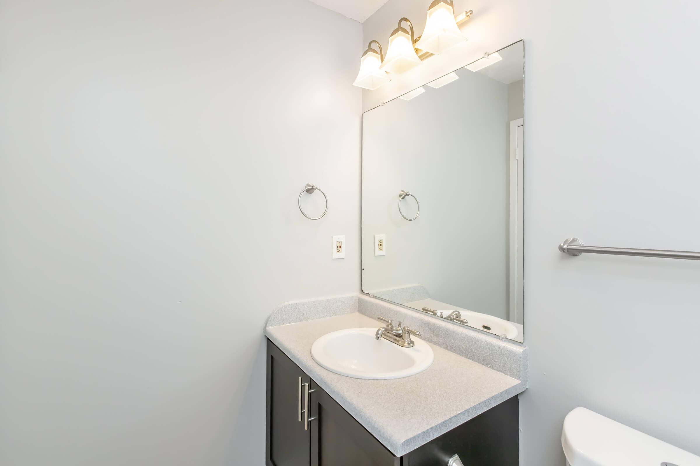 A clean, modern bathroom featuring a gray wall, a white countertop with a sink, and a large mirror above it. The sink area has a chrome faucet and a towel ring on the wall. To the right, there's a toilet, and a chrome towel bar is mounted on the wall. The overall style is simple and contemporary.