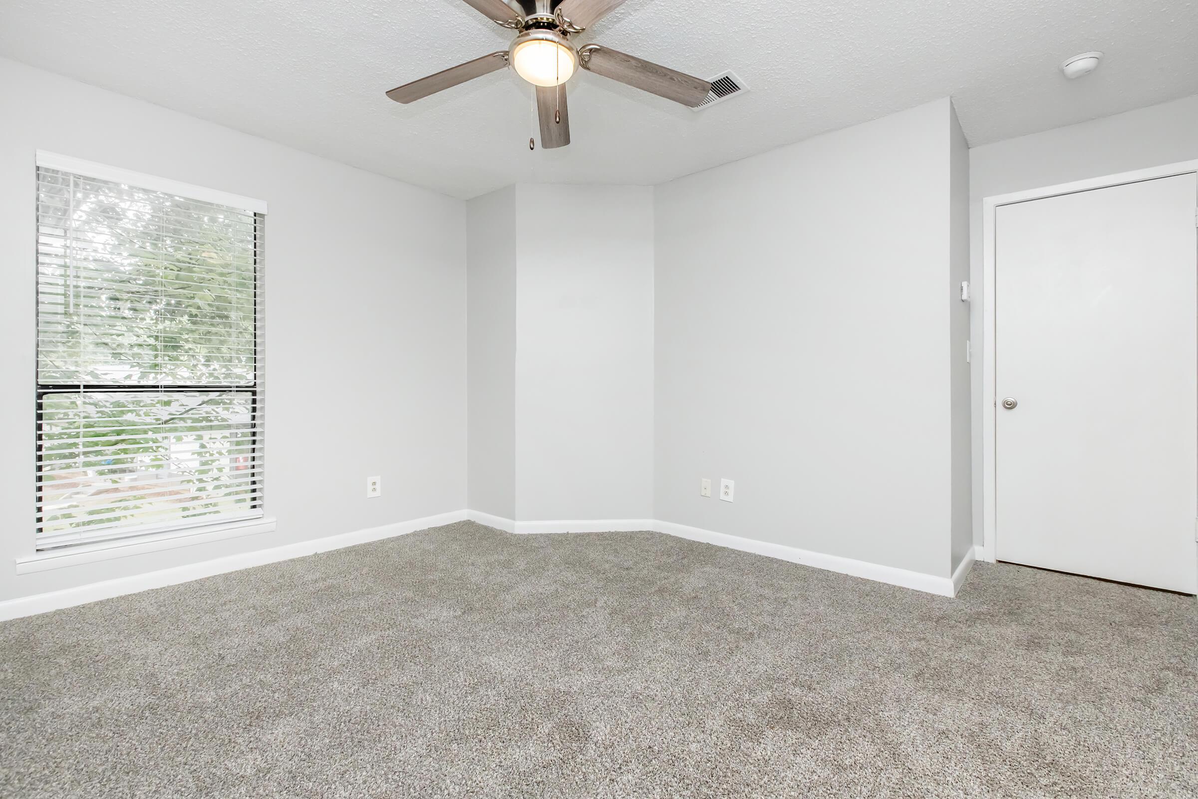 A vacant room with light gray walls and carpeted flooring. There is a ceiling fan with five blades, and a window with vertical blinds allowing natural light. A door is visible on the right, and the overall space appears clean and well-maintained, suitable for various uses.