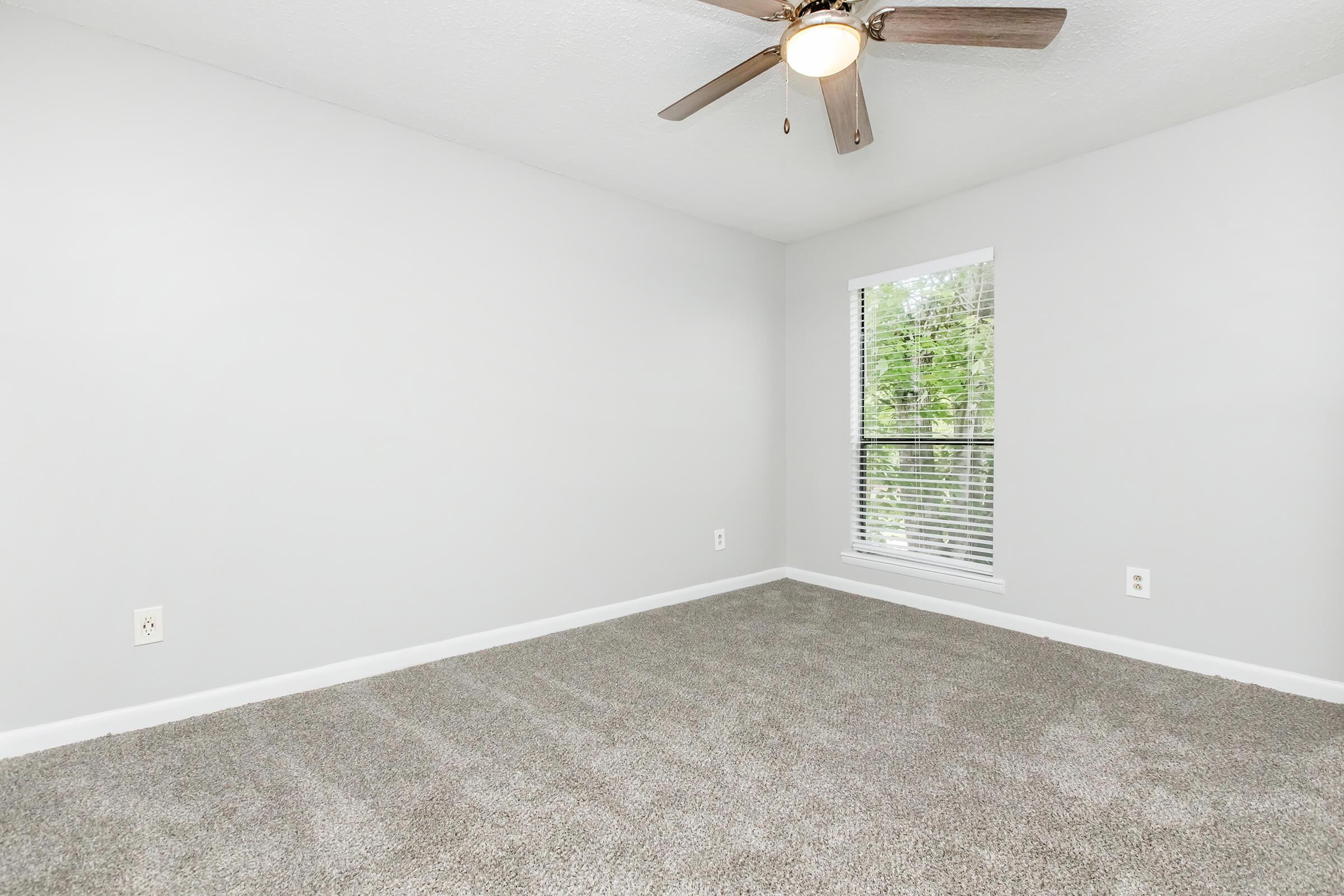 A sparse, empty bedroom with neutral gray walls and carpet. A ceiling fan is mounted on the ceiling, and there's a window with blinds letting in natural light. The room has a clean, modern feel with no furniture or decorations.