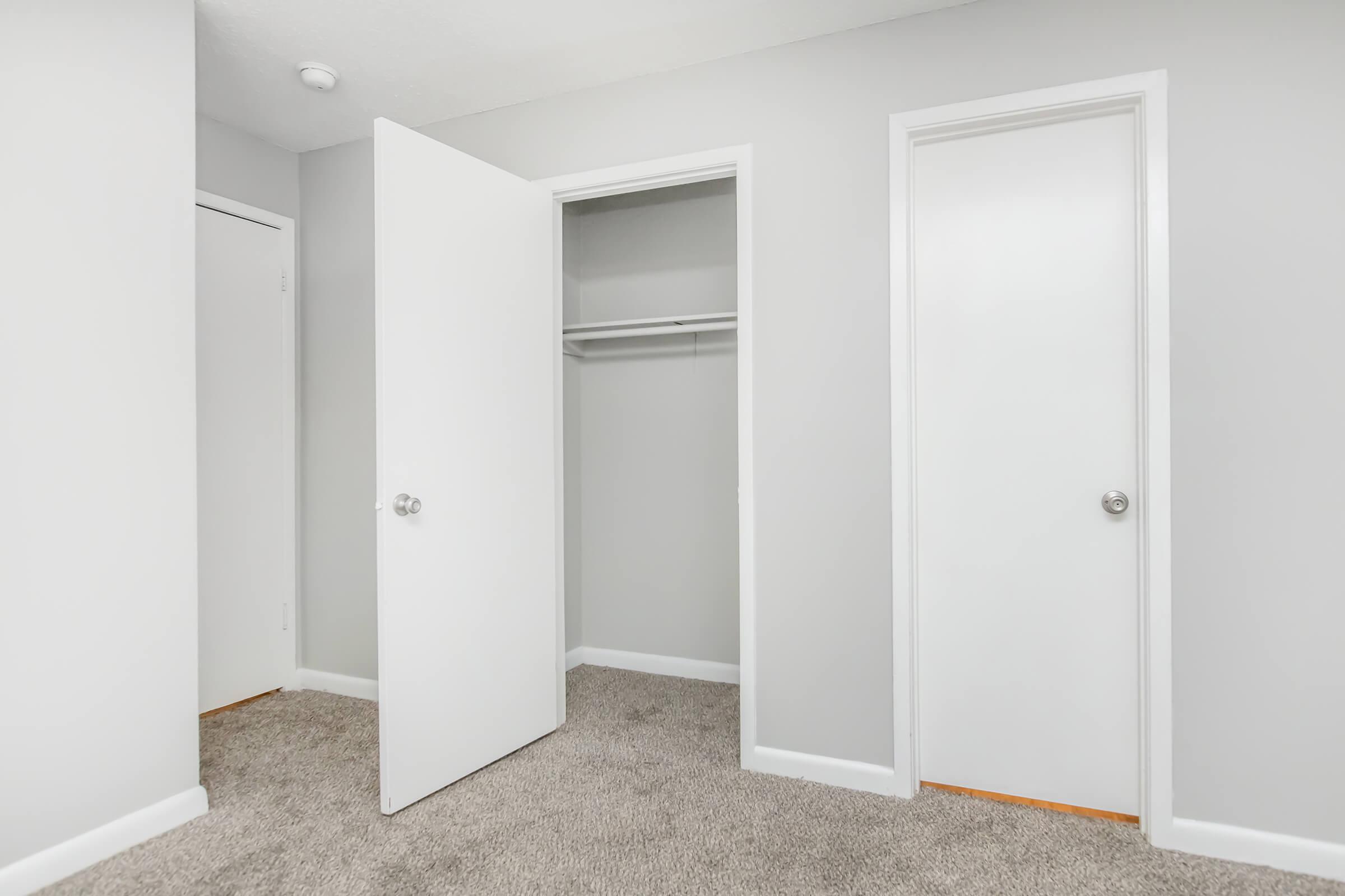 A small, empty room featuring light gray walls and a beige carpet. There are two closed white doors: one leads to a closet with a hanging rod, and the other appears to lead to another space. The room is well-lit, with natural light coming from an unseen source.