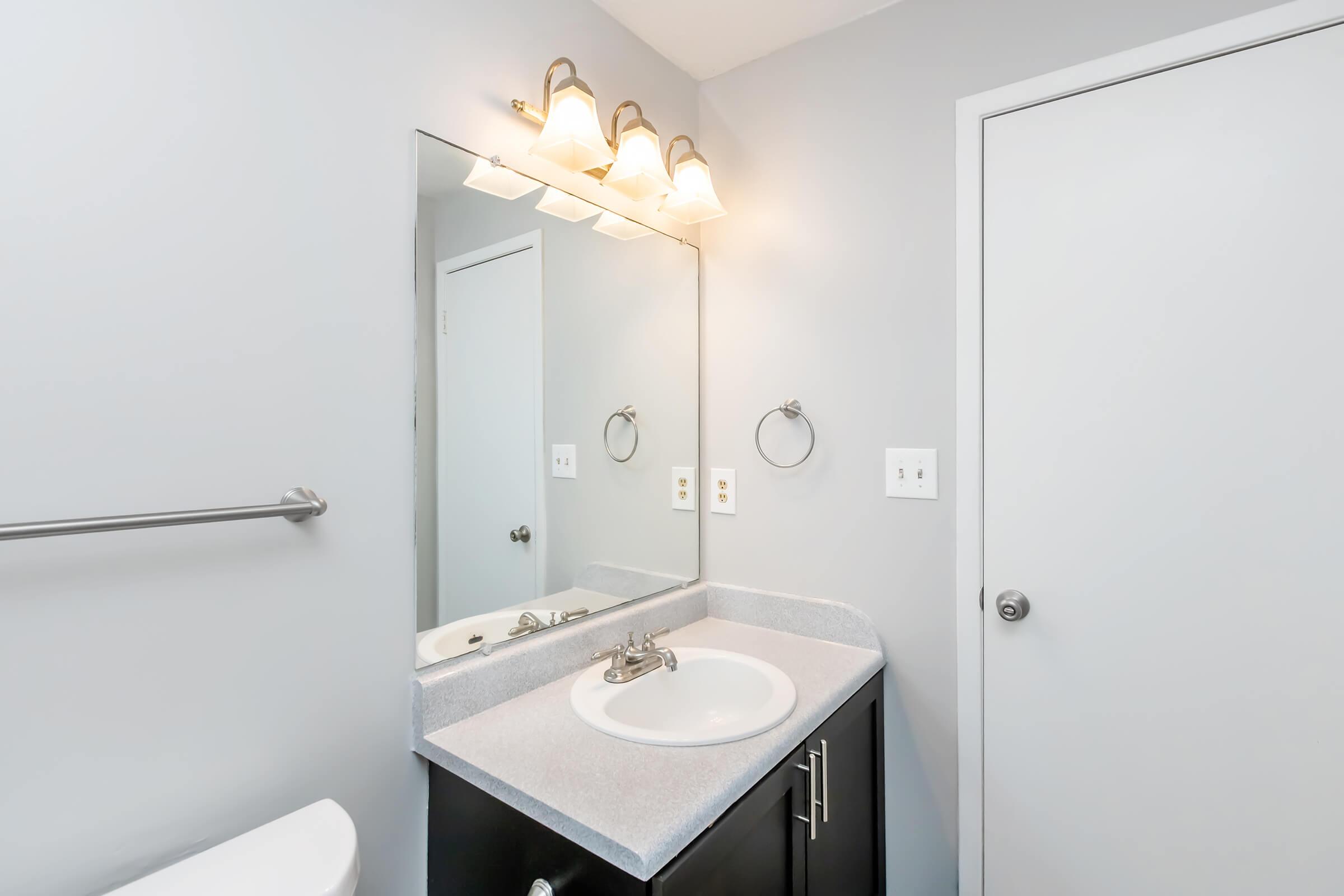 Bright and modern bathroom featuring a clean sink area with a white countertop, mirror, and three light fixtures above. A simple gray wall complements the design, and a door is visible on the right. A towel ring and grab bar add functionality to the space.