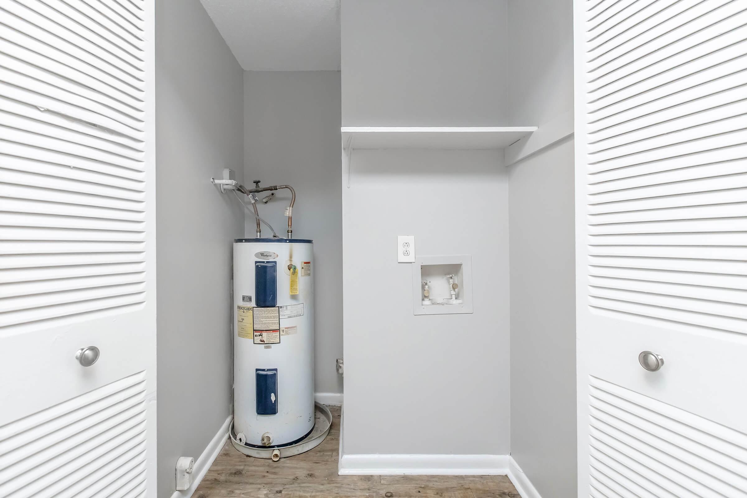 A small utility room featuring a water heater against a gray wall. There is a shelf above the heater, and a partially empty closet with louvered doors on either side. The floor is light-colored, and the space appears modern and clean.