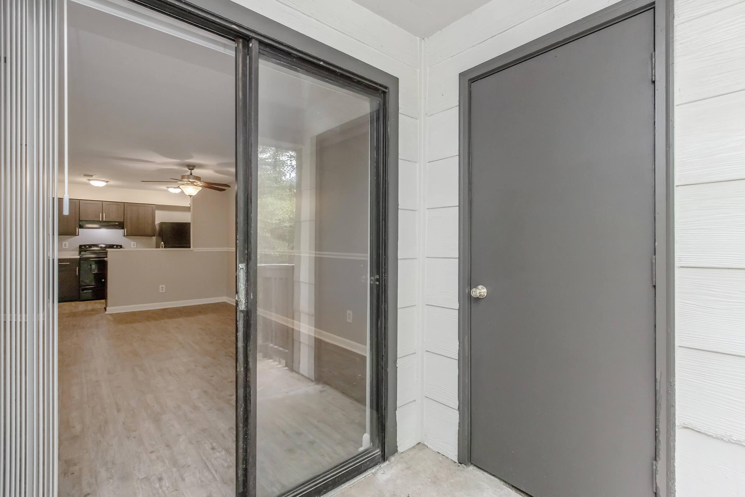 View of an entryway leading into a living space. The image shows a sliding glass door and a solid dark door, with a glimpse of the interior featuring a ceiling fan, light-colored walls, and a wooden floor. Natural light streams in, creating a welcoming atmosphere.