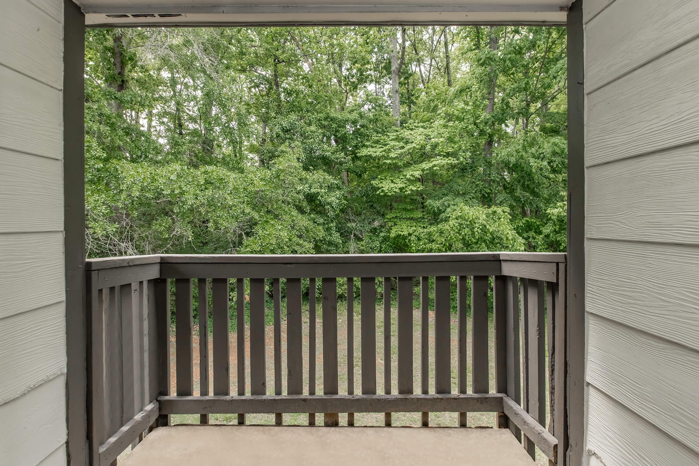 View from a balcony overlooking a lush, green wooded area. The railing is wooden, and the scene conveys a peaceful, natural setting with dense foliage in the background. Bright and inviting atmosphere with a sense of tranquility.