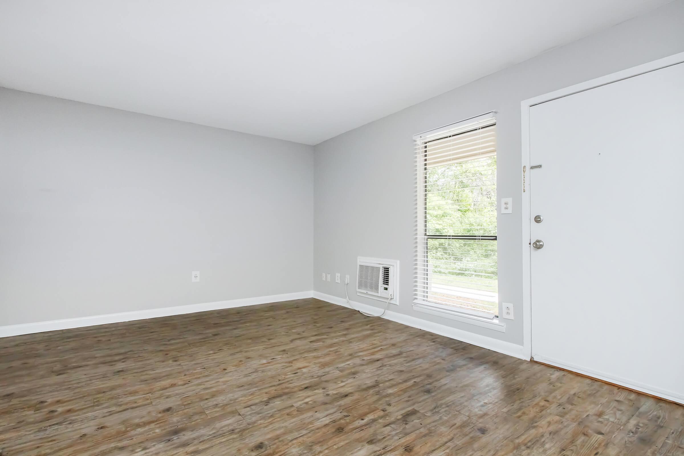 A spacious, empty room with light gray walls and a hardwood floor. There is a window with blinds providing natural light, and an air conditioning unit on the wall. The entrance door is on the right, featuring a simple design and a handle. The overall atmosphere is bright and inviting.