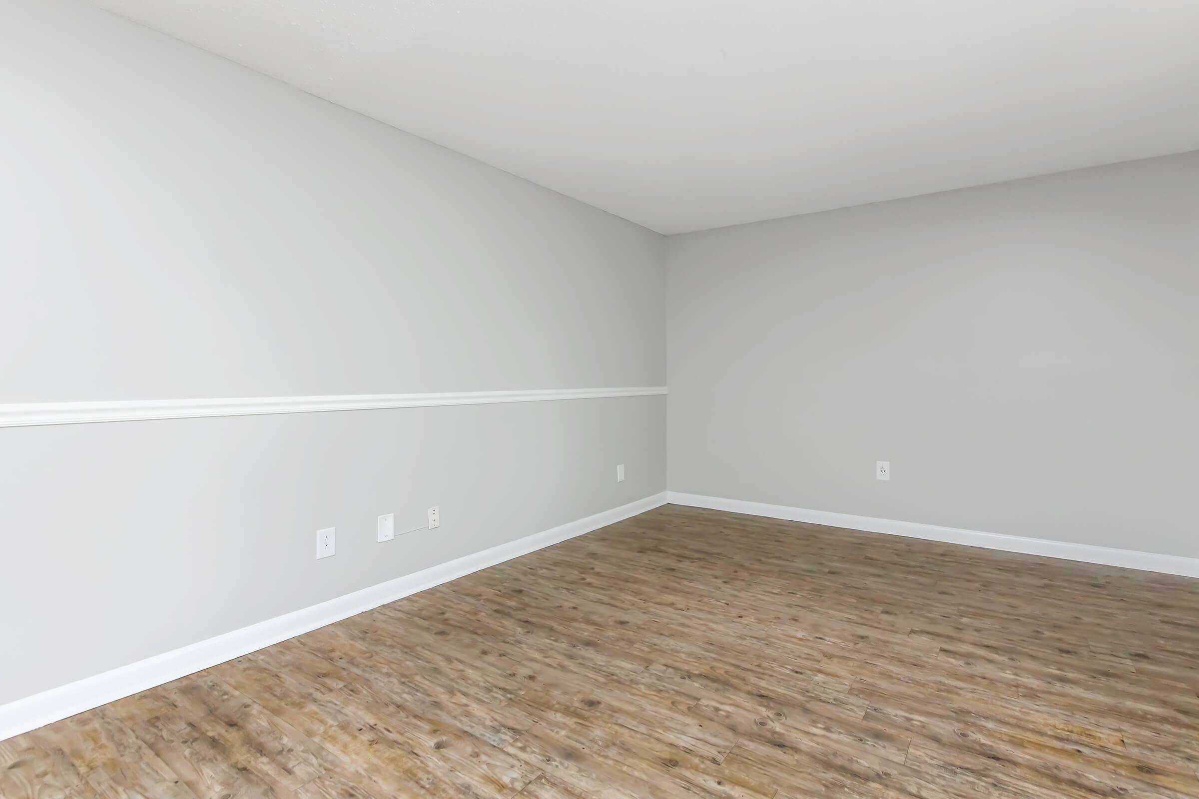 A spacious, empty room with light gray walls and a white chair rail. The floor features wood-like laminate in a warm tone. There are no furnishings or decorations, creating a blank canvas ideal for interior design or renovation. Natural light brightens the space.
