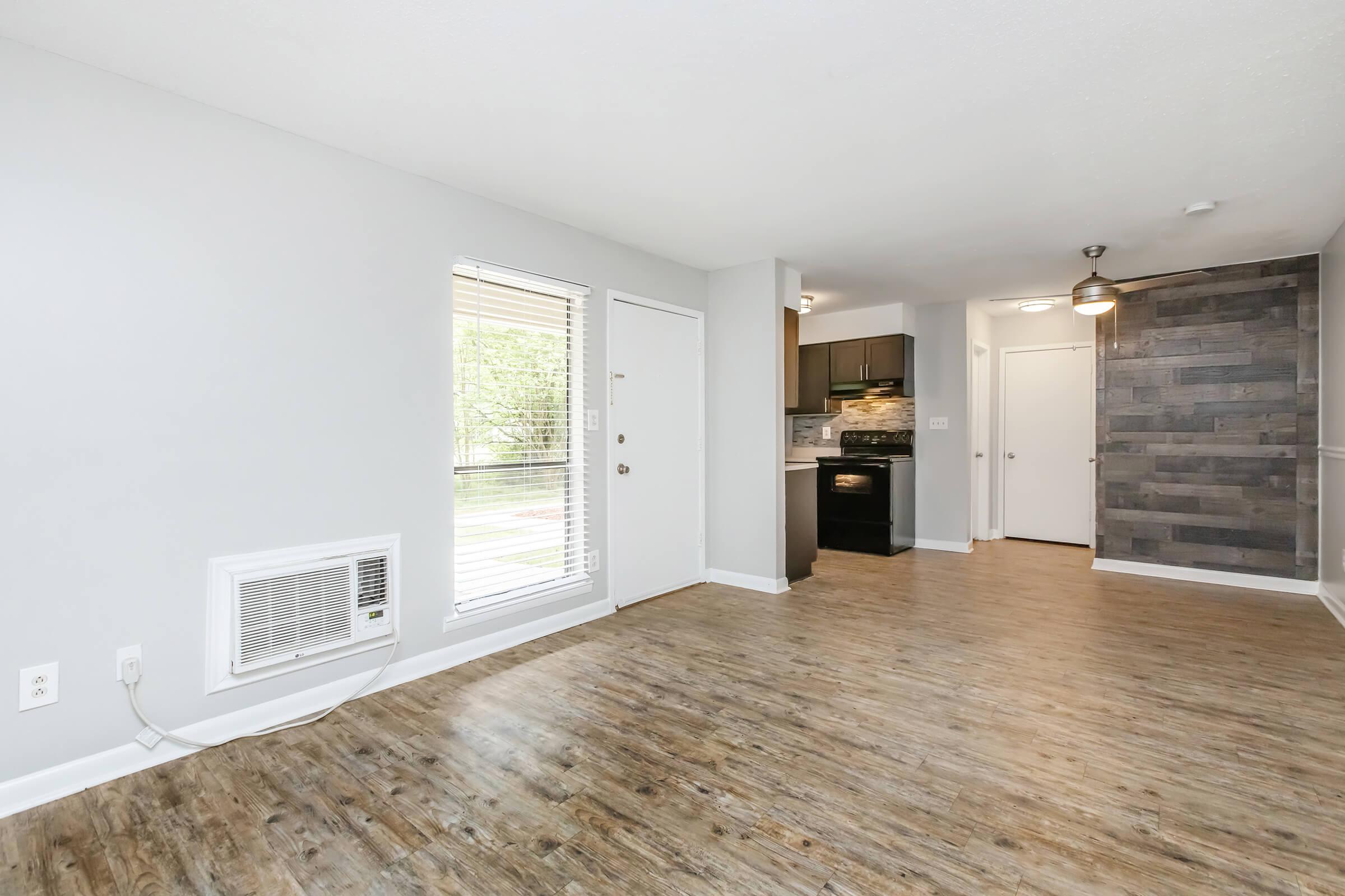 Spacious living area featuring hardwood-style flooring, a wall-mounted air conditioning unit, and large windows allowing natural light. The open layout includes a view of the kitchen with dark cabinetry and stainless steel appliances, creating a modern atmosphere.
