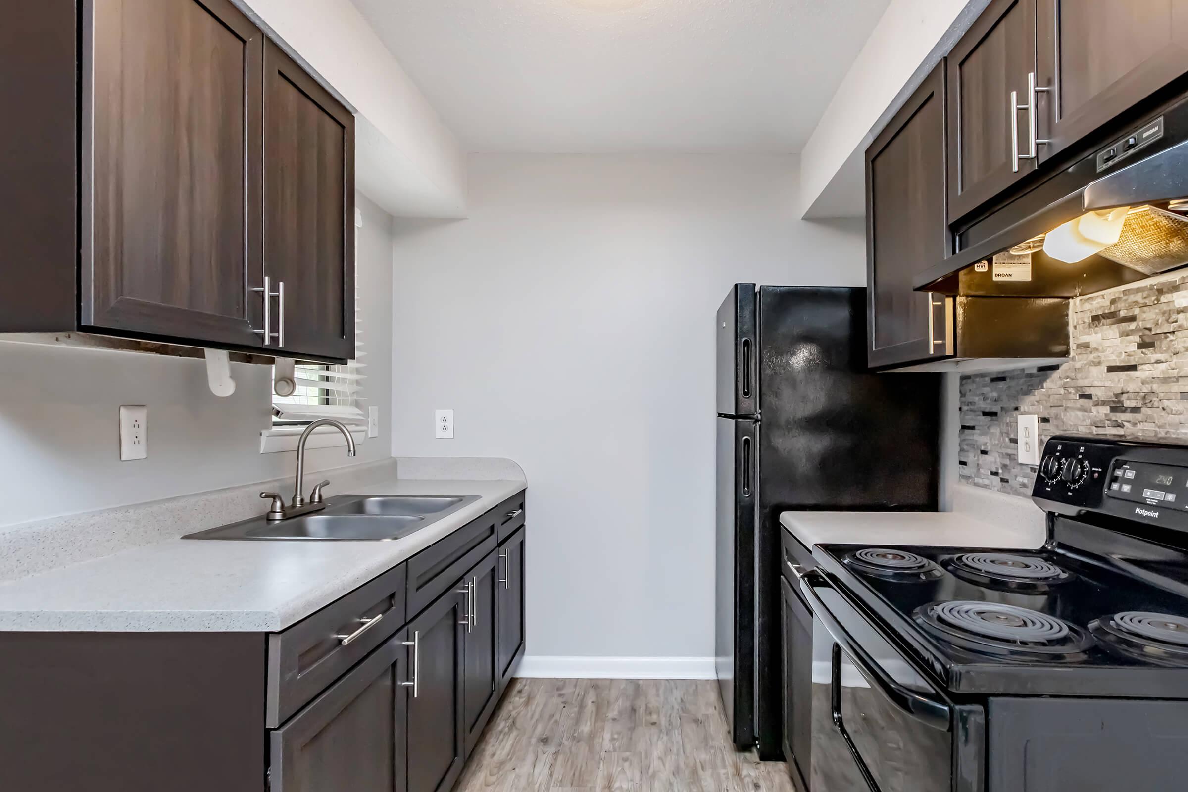 Modern kitchen featuring dark wood cabinetry, a stainless steel sink, and white countertops. Appliances include a black refrigerator and an electric stove with an oven. The backsplash consists of gray tiles, and natural light comes through a window, illuminating the space. The floor is light-colored laminate.