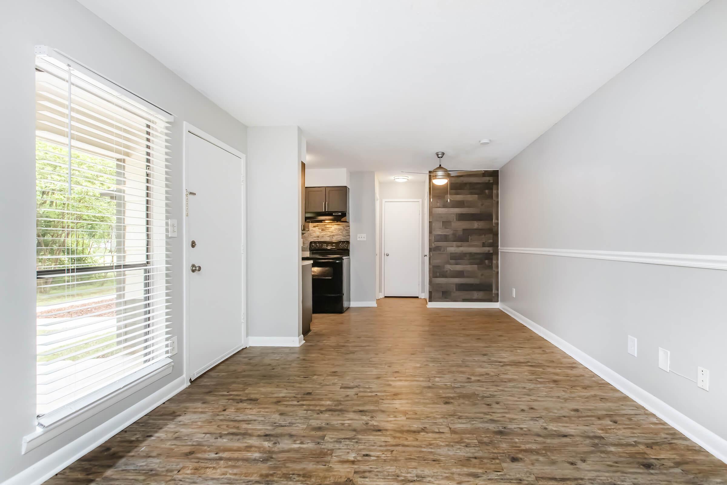 A bright, spacious living area featuring wood flooring, a large window with blinds, and an open layout. The kitchen is visible in the background with dark cabinets and modern appliances. The walls are painted in neutral tones, and there's a stylish accent wall.