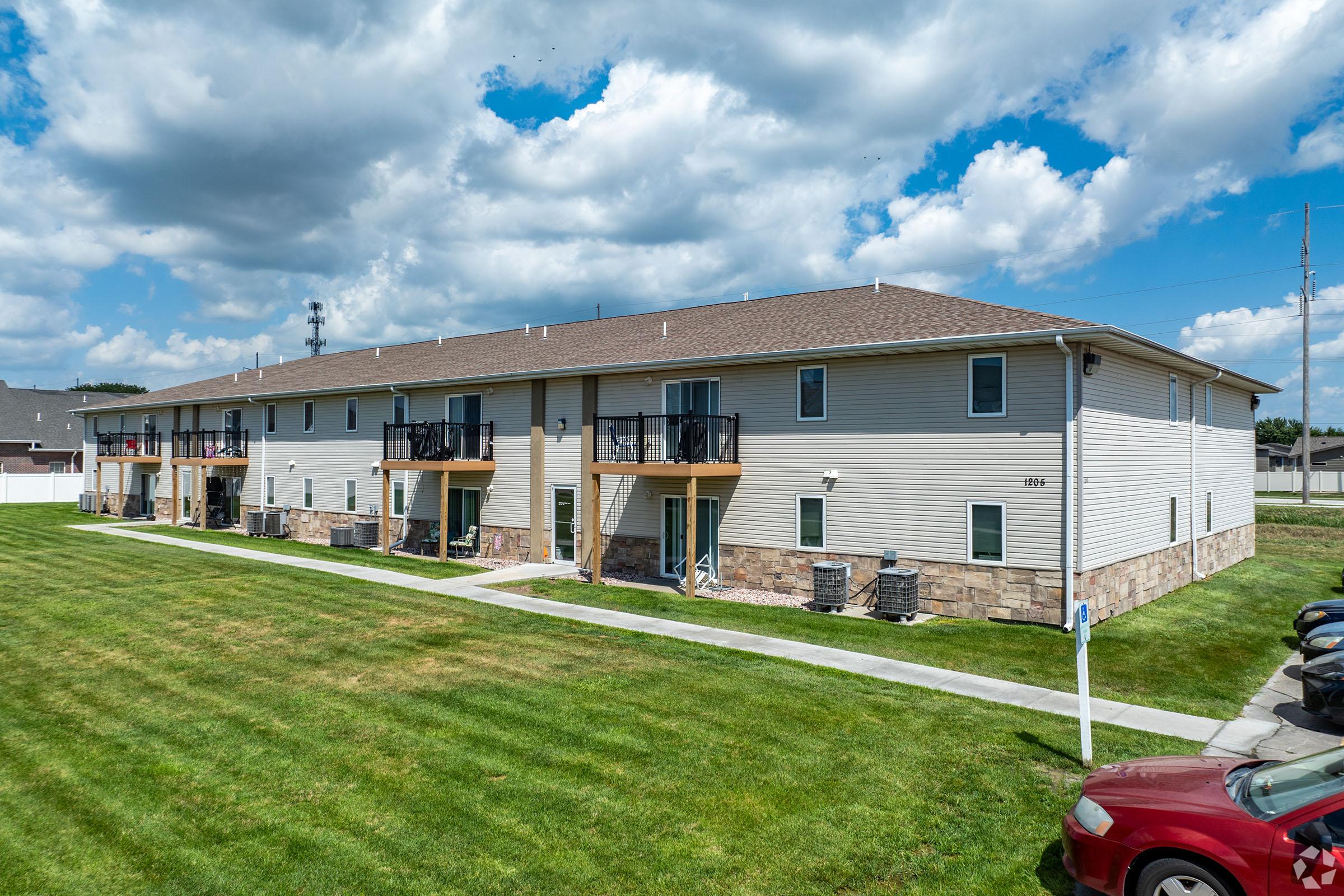 A two-story apartment building with beige siding and stone accents, featuring multiple balconies. The grassy area in front has a sidewalk and several parking spaces. The sky is partly cloudy, and there are utility poles in the background.