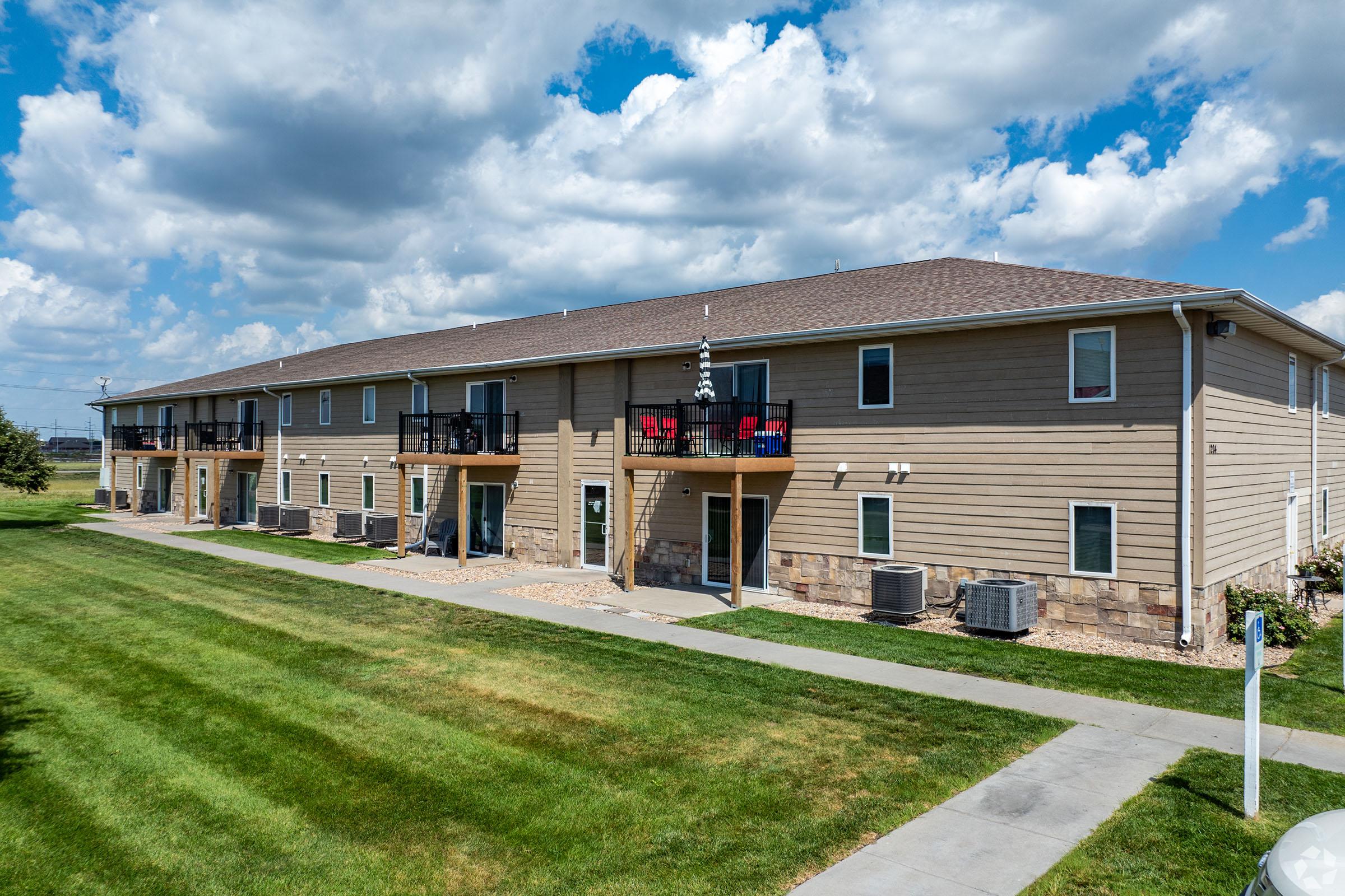 A two-story apartment building with a light brown exterior, featuring balconies with red chairs. The grassy area in front is well-kept, with a sidewalk leading to the entrance. In the background, there are blue skies with scattered clouds. Air conditioning units are visible along the side of the building.